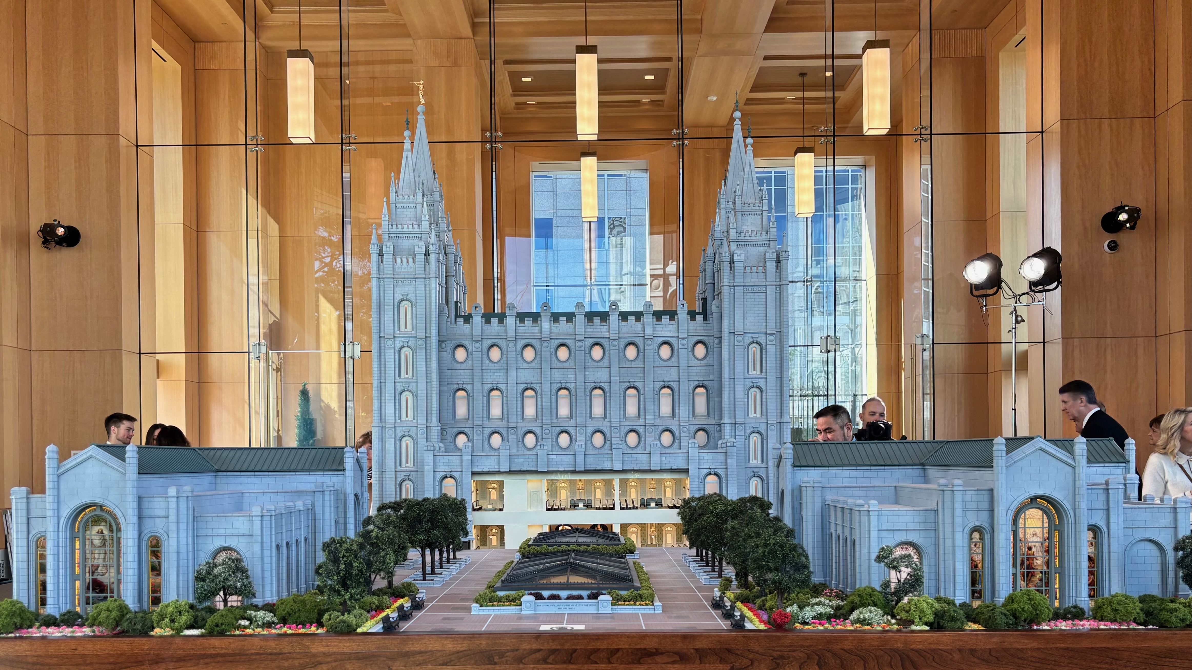 Intricate blue-gray castle model with twin spires and arched windows sits inside a glass-walled showroom. A landscaped courtyard with trees and a central feature surrounds the display.