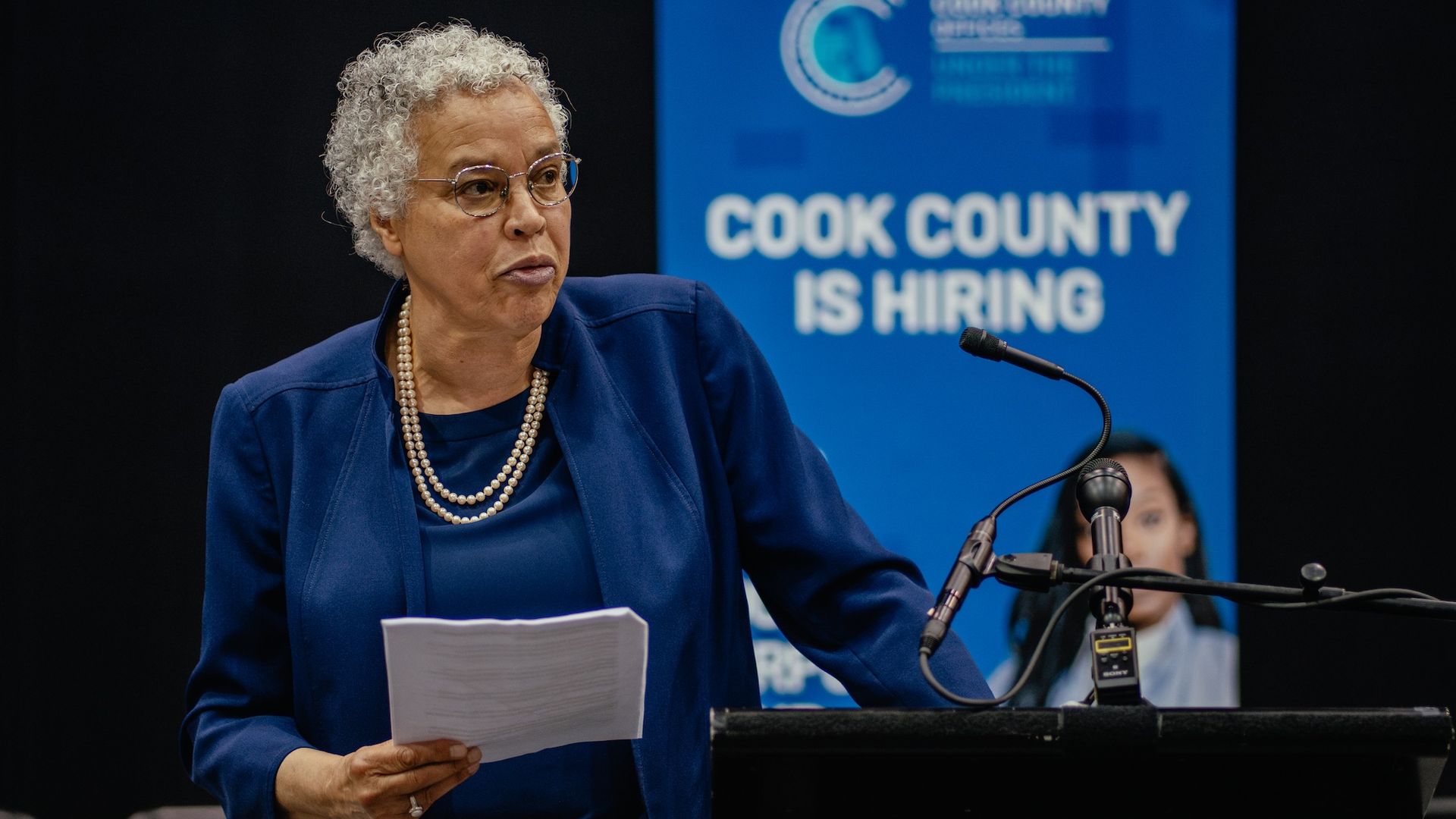 Cook County president Toni Preckwinkle in a blue suit and white pearls holding a white paper in front of a blue banner reading "Cook County is Hiring"