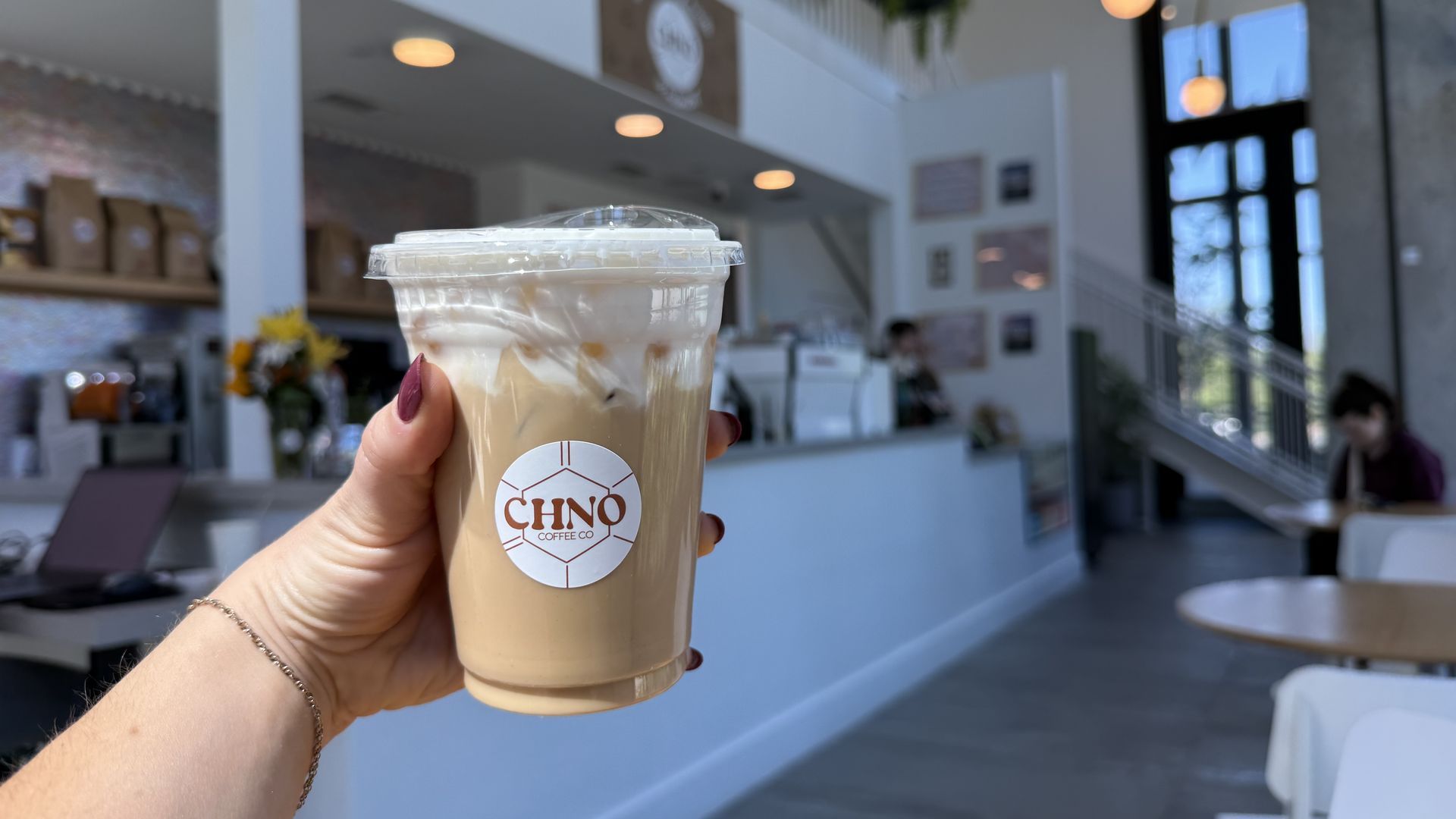 Hand holding iced coffee in a clear cup with whipped cream, CHINO Coffee Co. logo, inside a bright cafe with white walls, warm lights, shelves, and a person at a table in the background.
