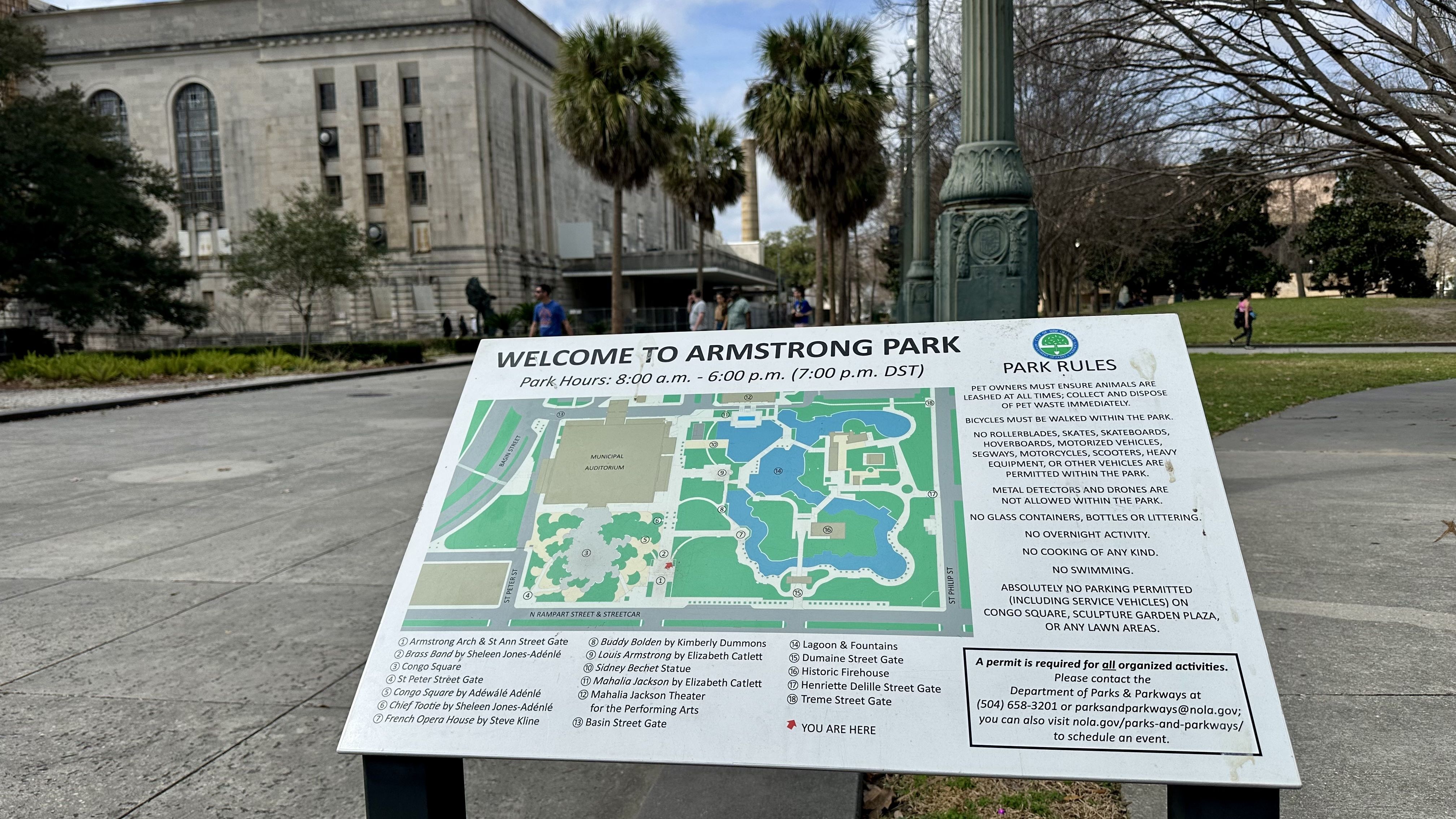 Map and park rules sign at Armstrong Park showing hours, landmarks, and regulations against certain vehicles, glass containers, overnight activity, and parking. Trees and building in background.