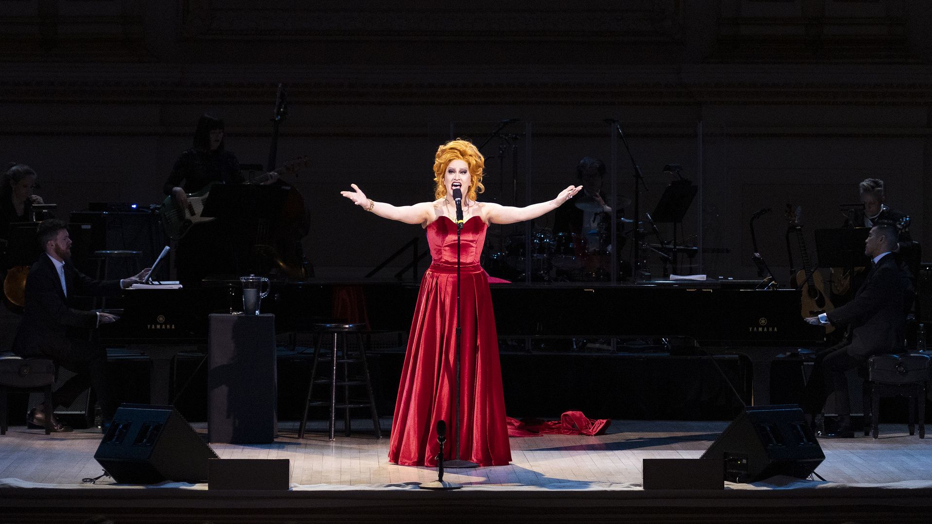 Performer in a long red gown with red hair sings center stage with arms extended, flanked by musicians playing instruments under dim lighting in a concert hall.
