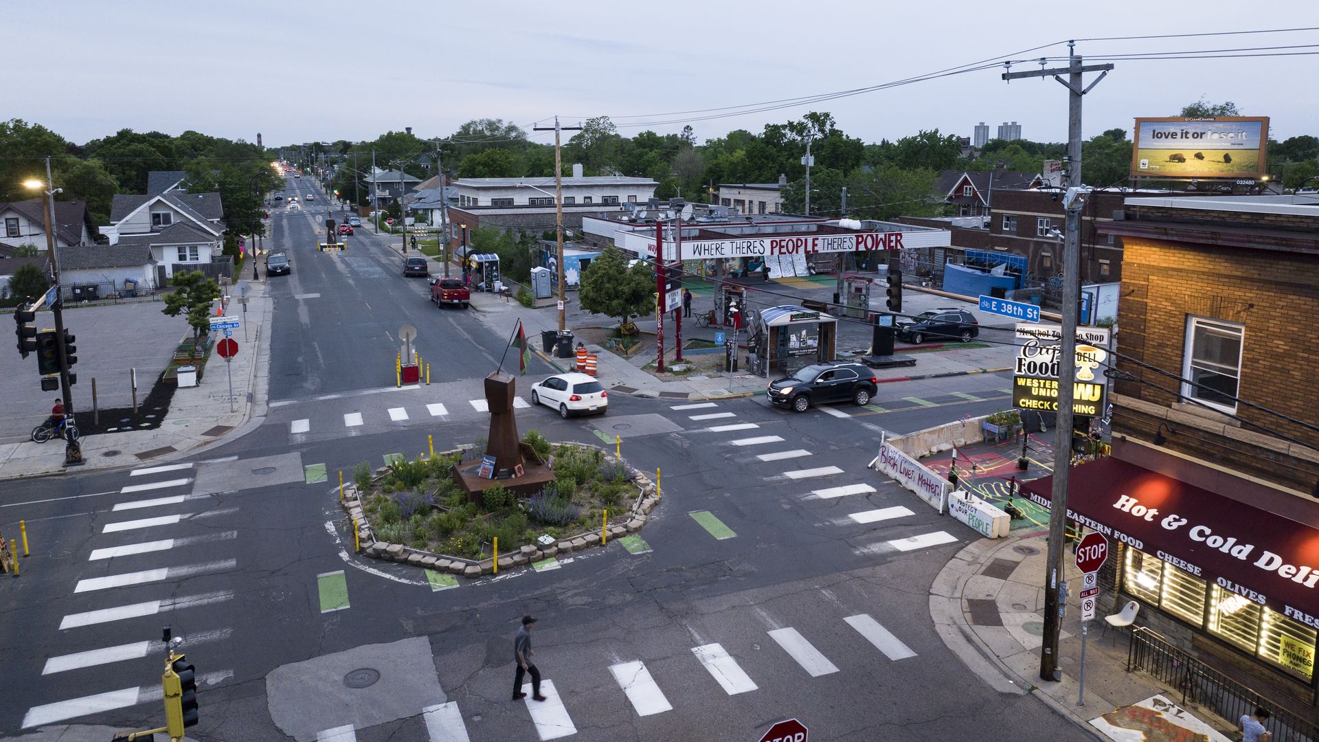 An overhead aerial shot of an intersection with a makeshift traffic circle and a sculpture near a convenience store