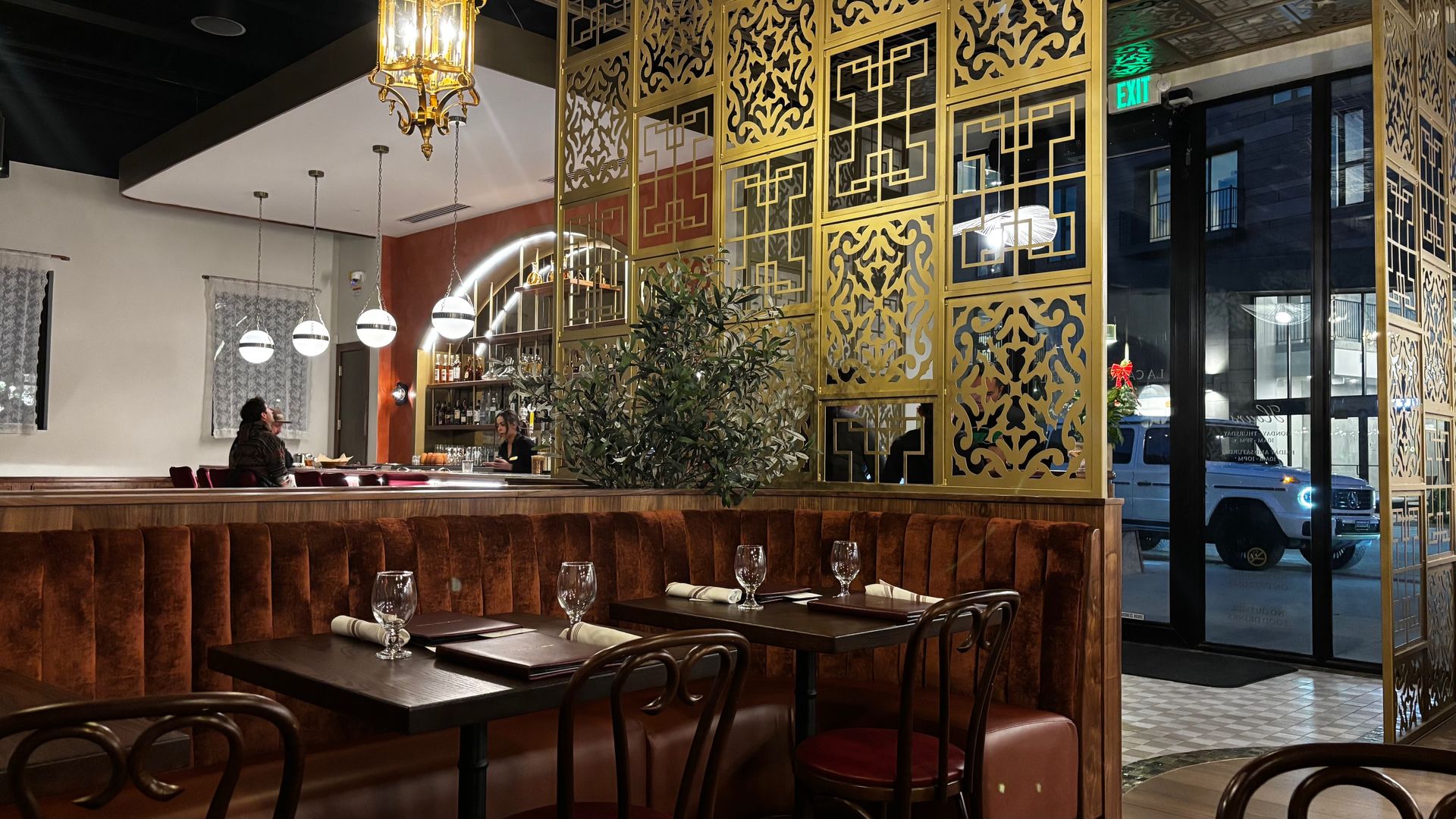 Cozy restaurant interior with brown velvet booths, dark wood tables set with glasses and napkins, gold ornate dividers, round hanging lights, and a bar with bottles behind.