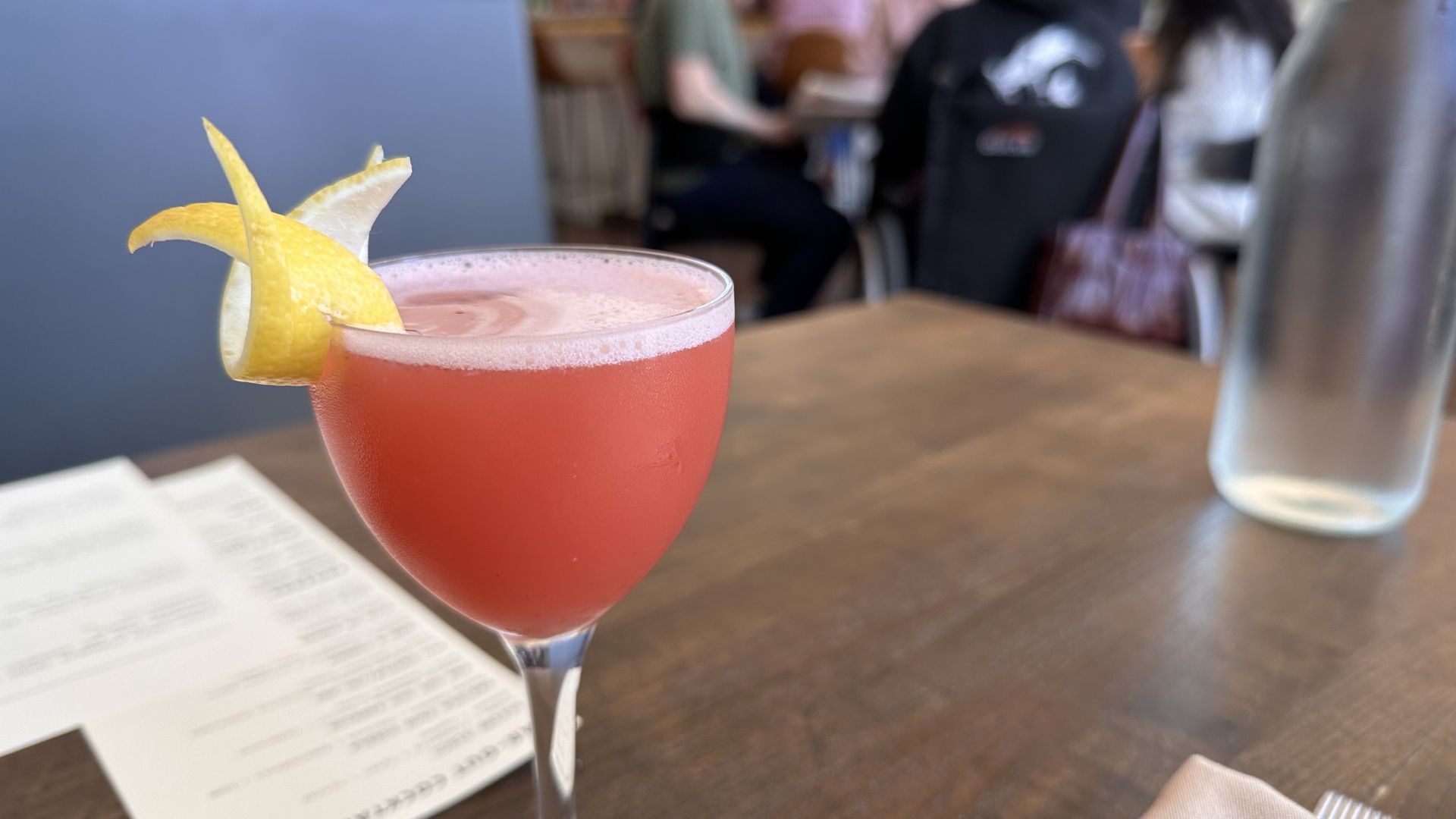 A pink cocktail with a lemon twist sits in a stemmed glass on a wooden table, with a blurred cafe scene of people and a water bottle in the background.