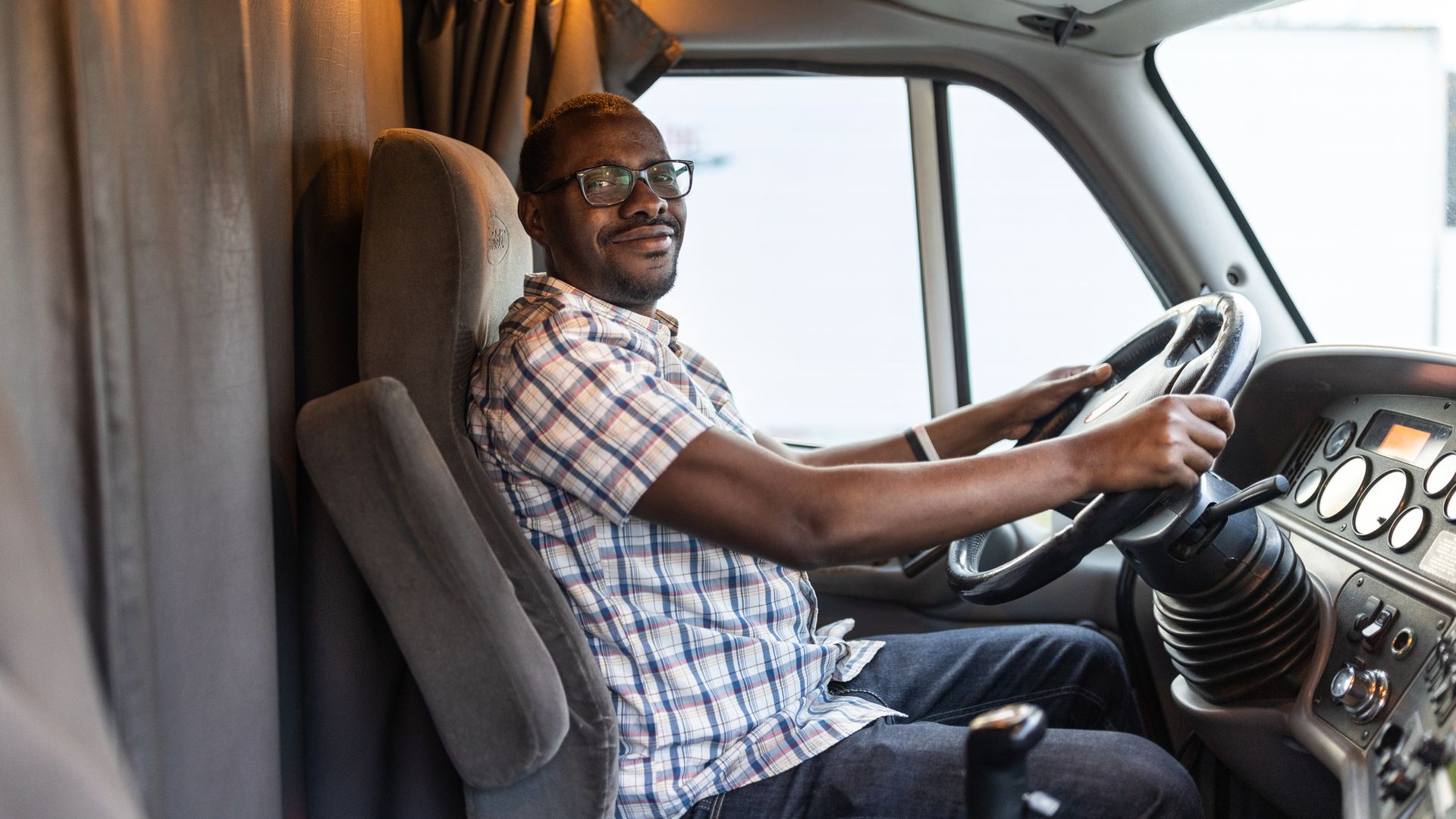 A man faces turns to face the camera while sitting in the driver's seat of a semi truck and holds the steering wheel. 