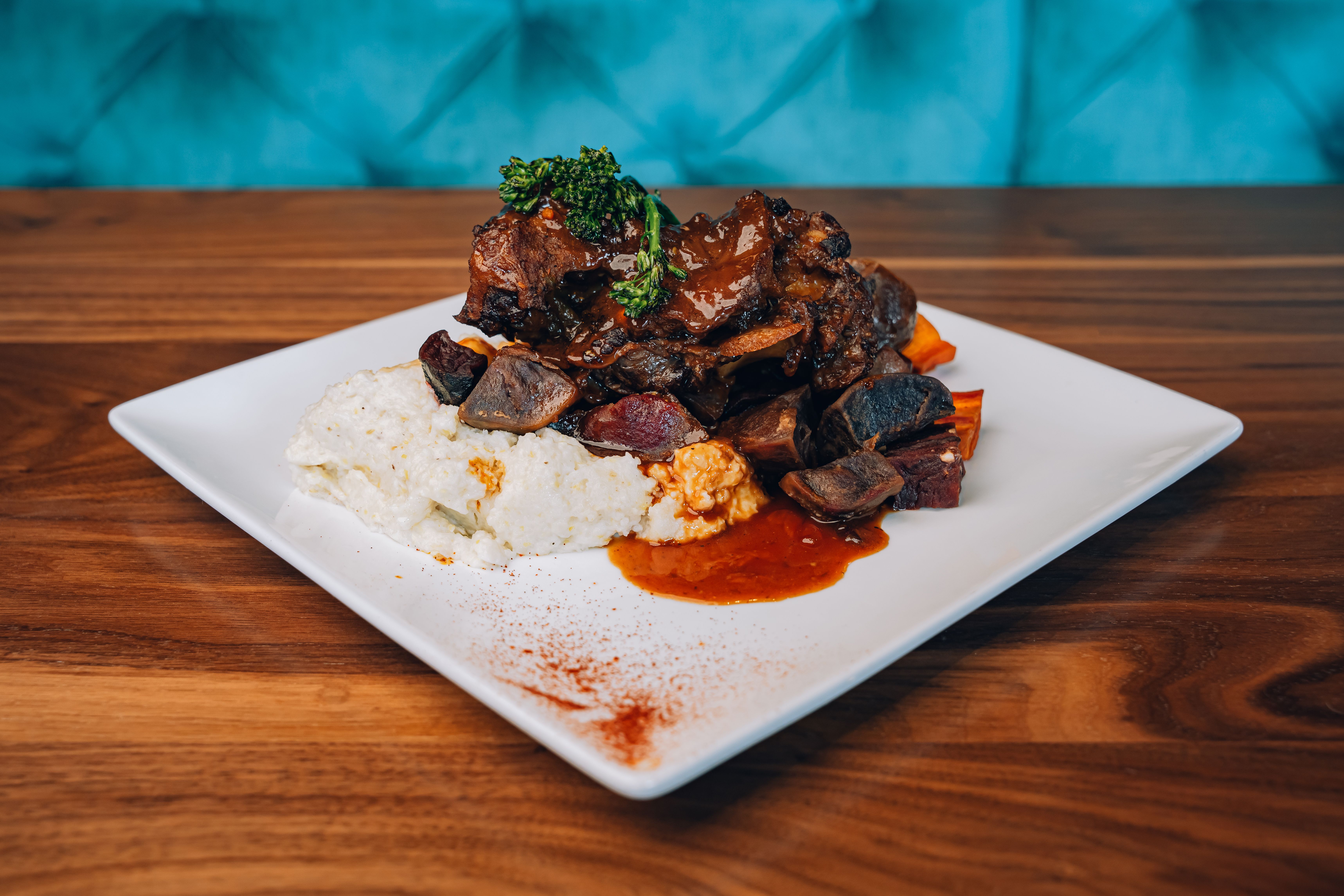 The oxtail and grits: Molasses-braised beef oxtails, stewed vegetables and coconut grits are served on a white square plate.