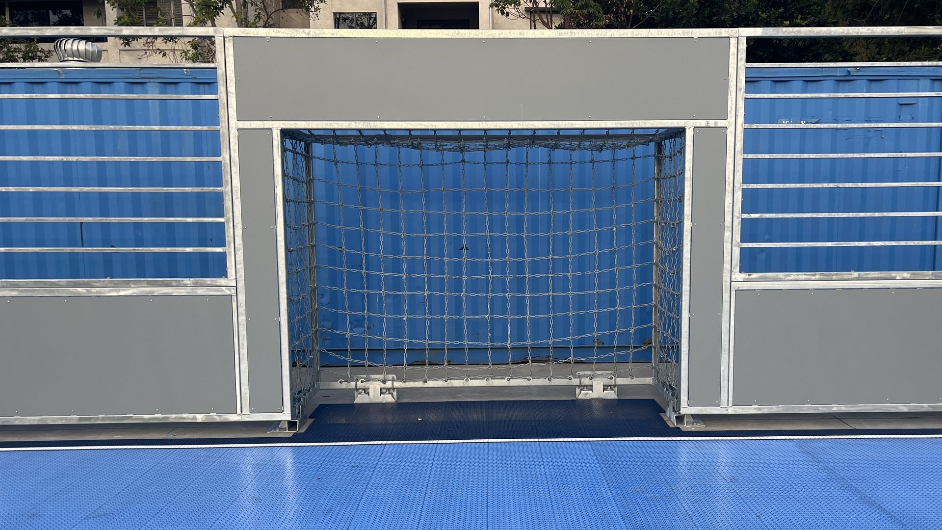 A small goal with a chain net on a soccer pitch with grey walls and blue flooring.