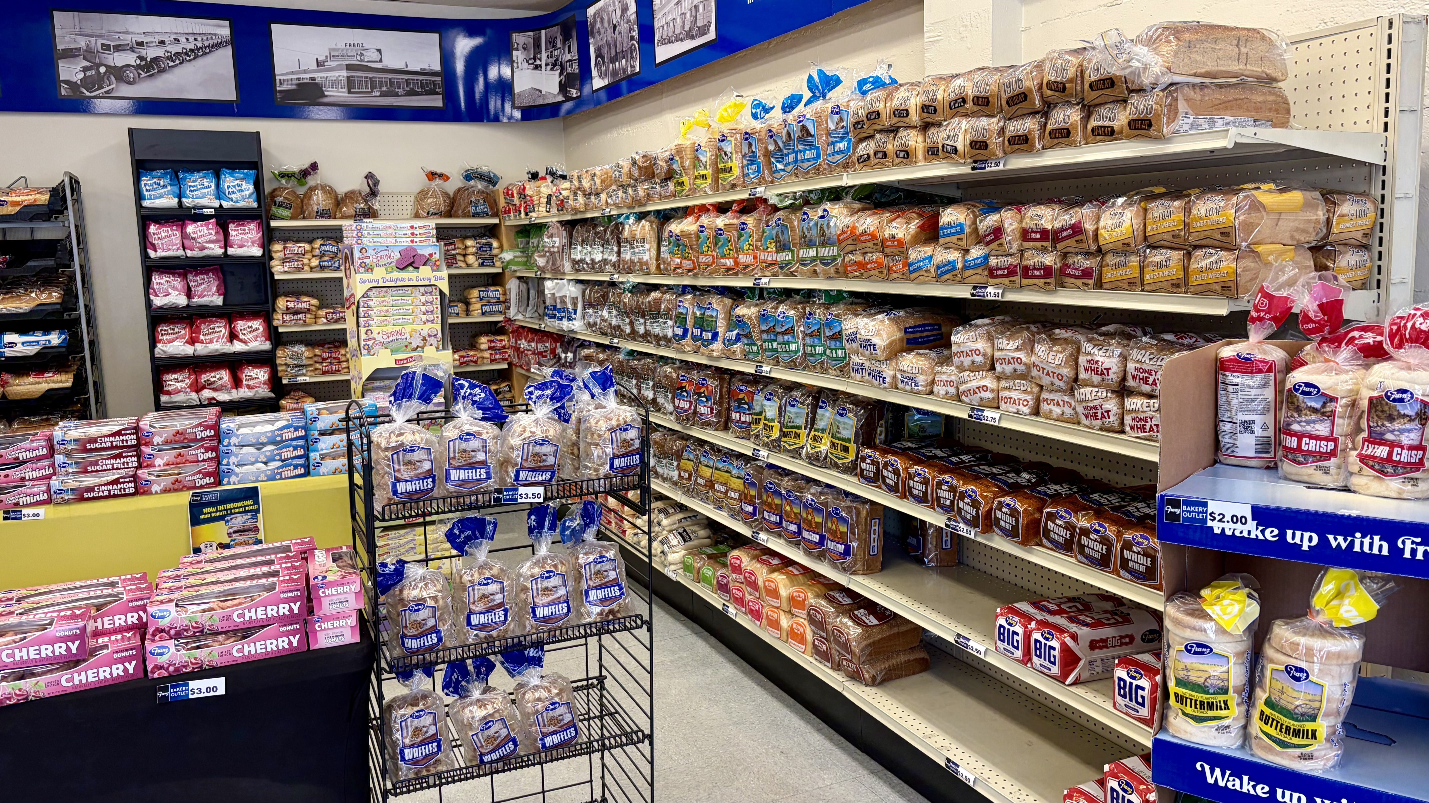 Bright grocery bakery aisle with long shelves of assorted loaves and bagged bread, a cart of waffles in front, pink and purple pastry boxes on the left, blue signage overhead.
