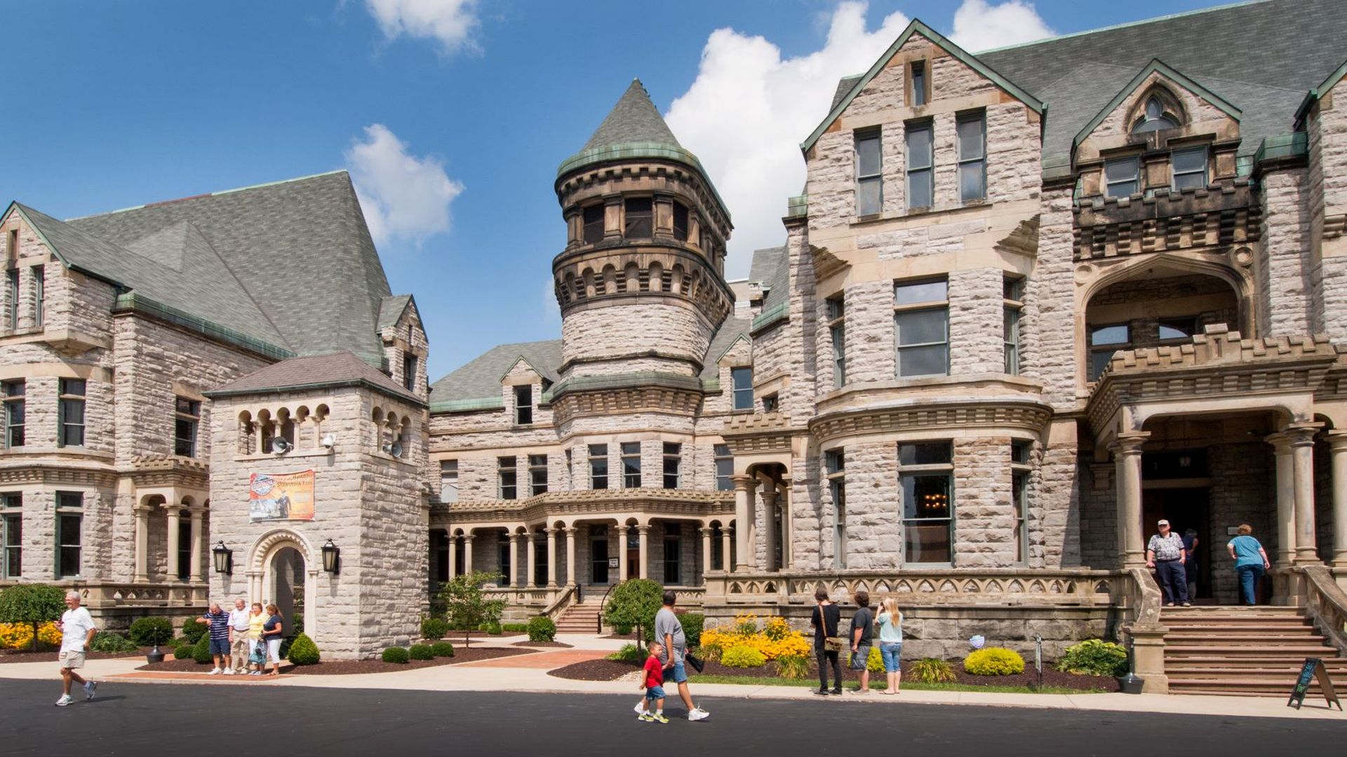 Castle-style stone building with a round turret and green roof, arched windows, and ornate stonework. People walk on a sunny plaza in front beneath a bright blue sky.