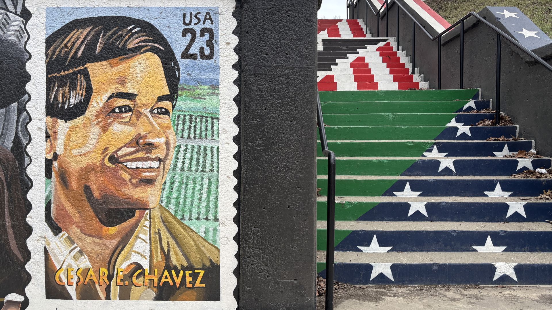 Vibrant mural of Cesar Chavez on the left in a stamp-like design with "USA 23" and "CESAR E CHAVEZ". A dark pillar divides from a right stairway painted green and blue with white stars and red stripes, with black rails.