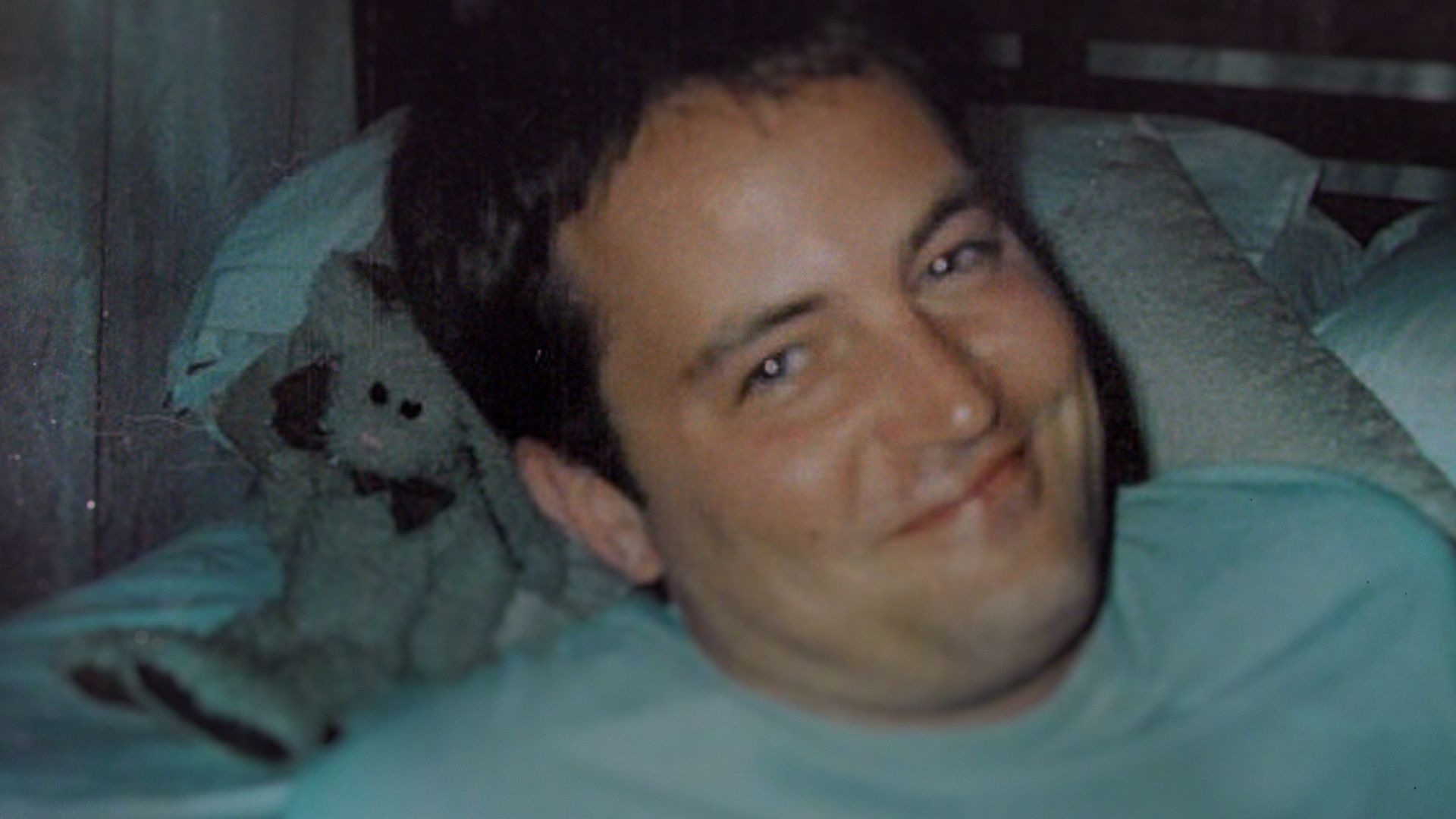 A young Matthew Perry lies in bed, head resting on teal blankets and a teal shirt. He smiles softly at the camera in a dim, casual bedroom setting.