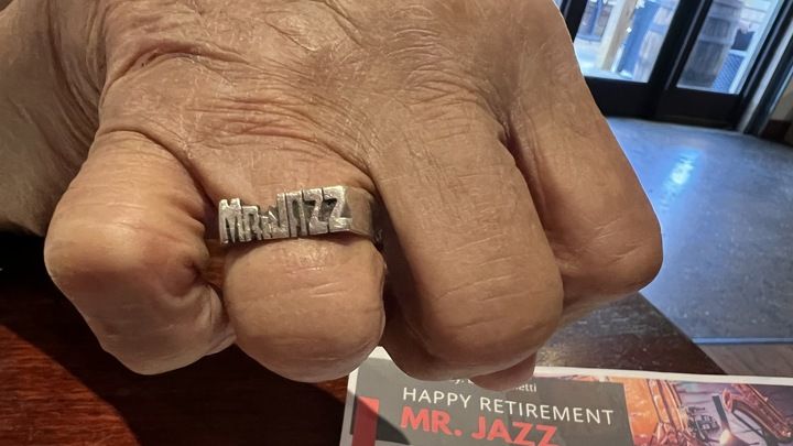 Close-up of a hand wearing a silver ring engraved with "MR. JAZZ" over a card that says "Happy Retirement MR. JAZZ" with a background of an indoor setting with glass doors.