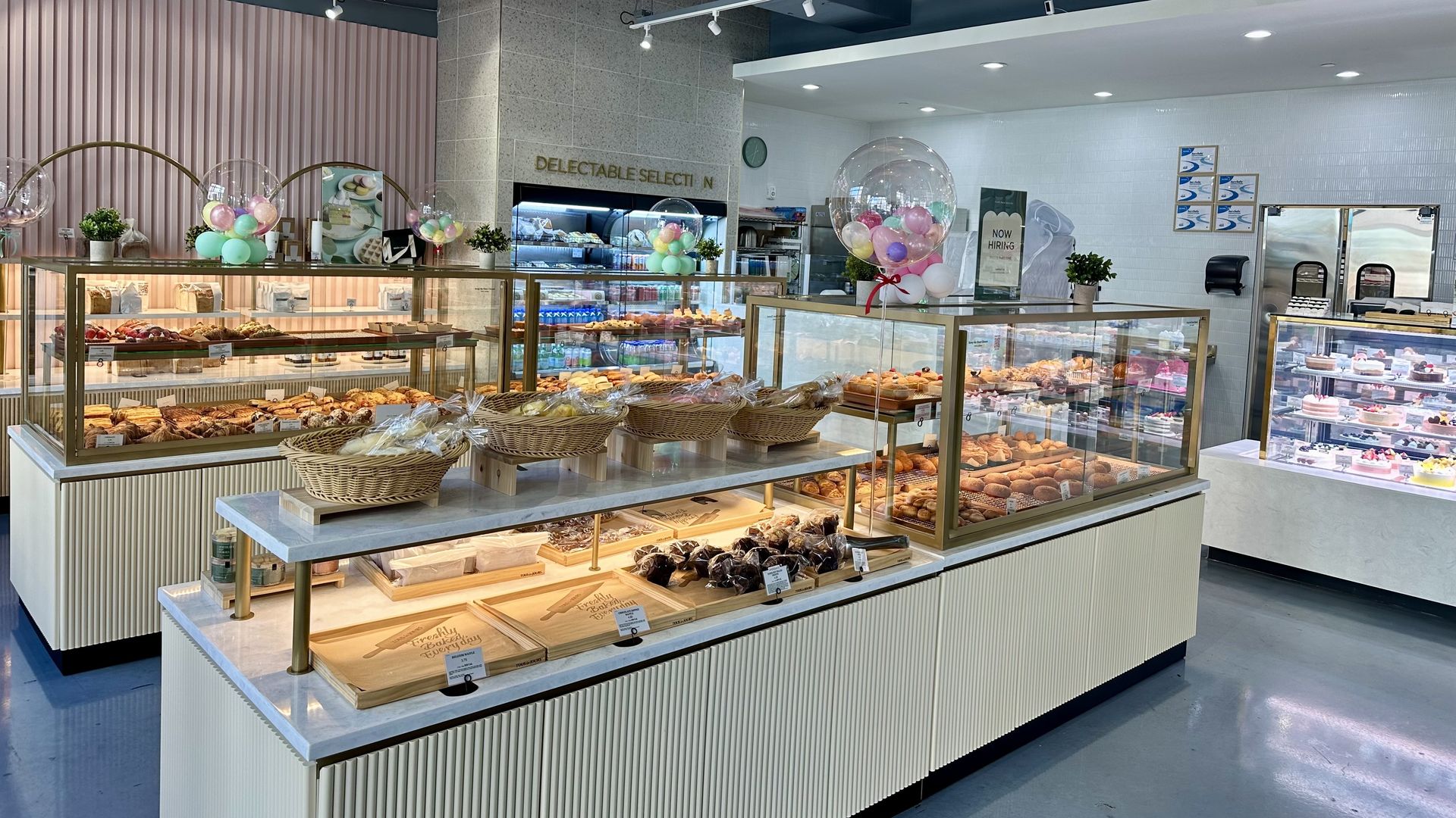 Bright bakery interior with glass display cases filled with various breads, pastries, and cakes. Pastel balloons and small potted plants decorate counters under exposed blue ceiling with track lighting.