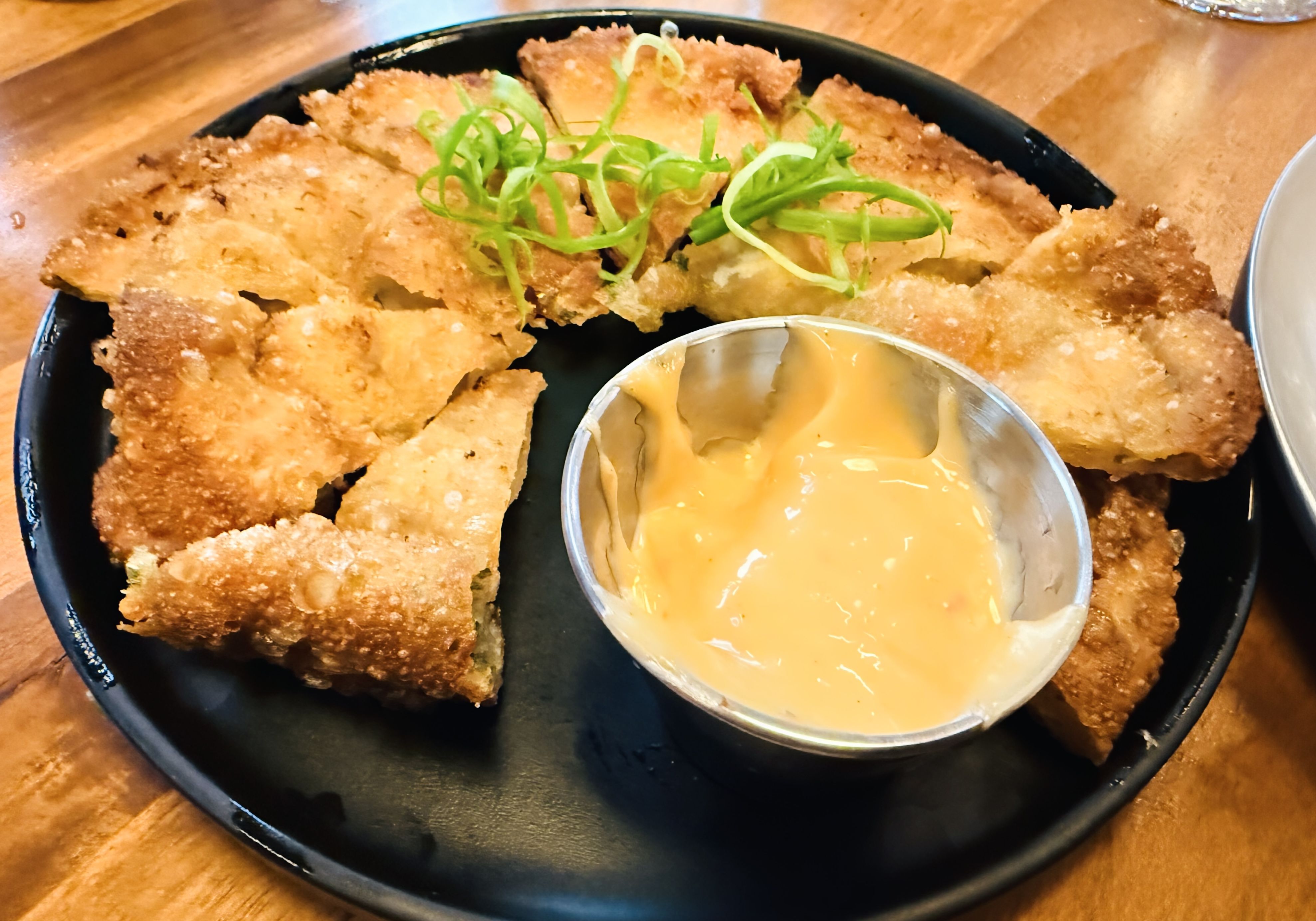 Golden-brown, crispy fried flat pieces on a black plate, topped with bright green onion curls; a small metal cup of pale yellow dipping sauce sits nearby on a wooden table.