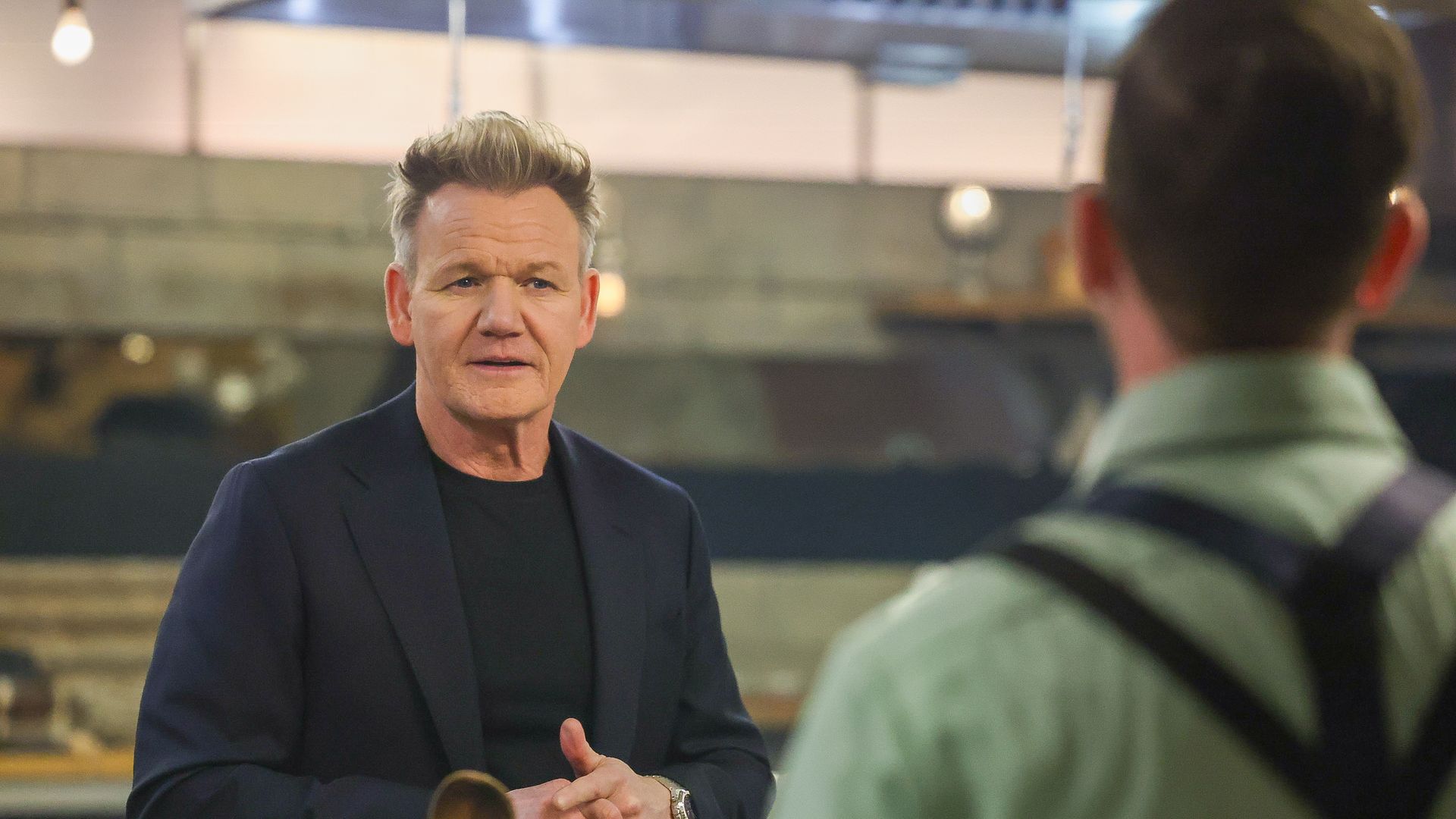 Gordon Ramsay, wearing a black shirt and dark blazer, speaks to a contestant in a kitchen setting for Next Level Chef.