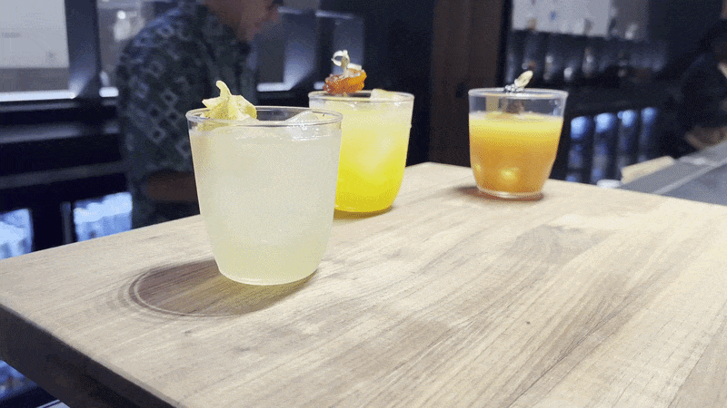 A video pans away from three cocktails set on a wooden cutting board. Behind the bar, a man looks down.