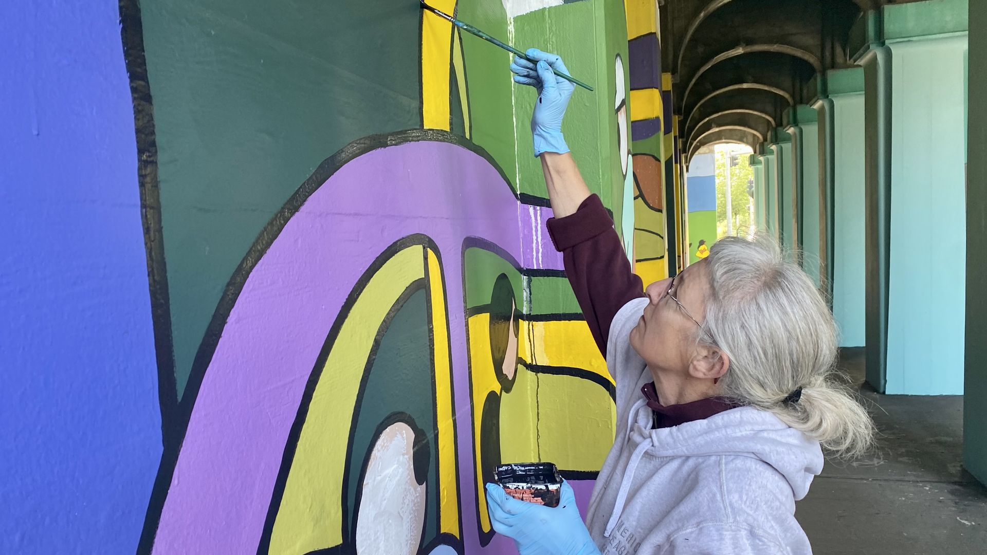 A woman reaches high above her head with a paintbrush to touch up a mural on a cement wall, with columns of an overpass behind her.