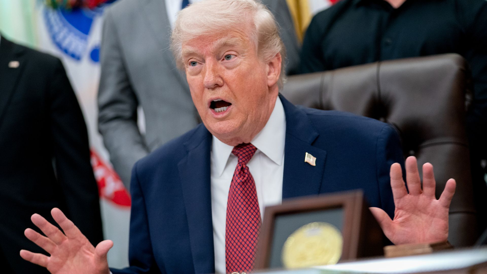 A President Trump with light blond hair sits at a desk, mouth open as if speaking, with both hands raised. He wears a dark blue suit, white shirt, red patterned tie, and a small flag pin.