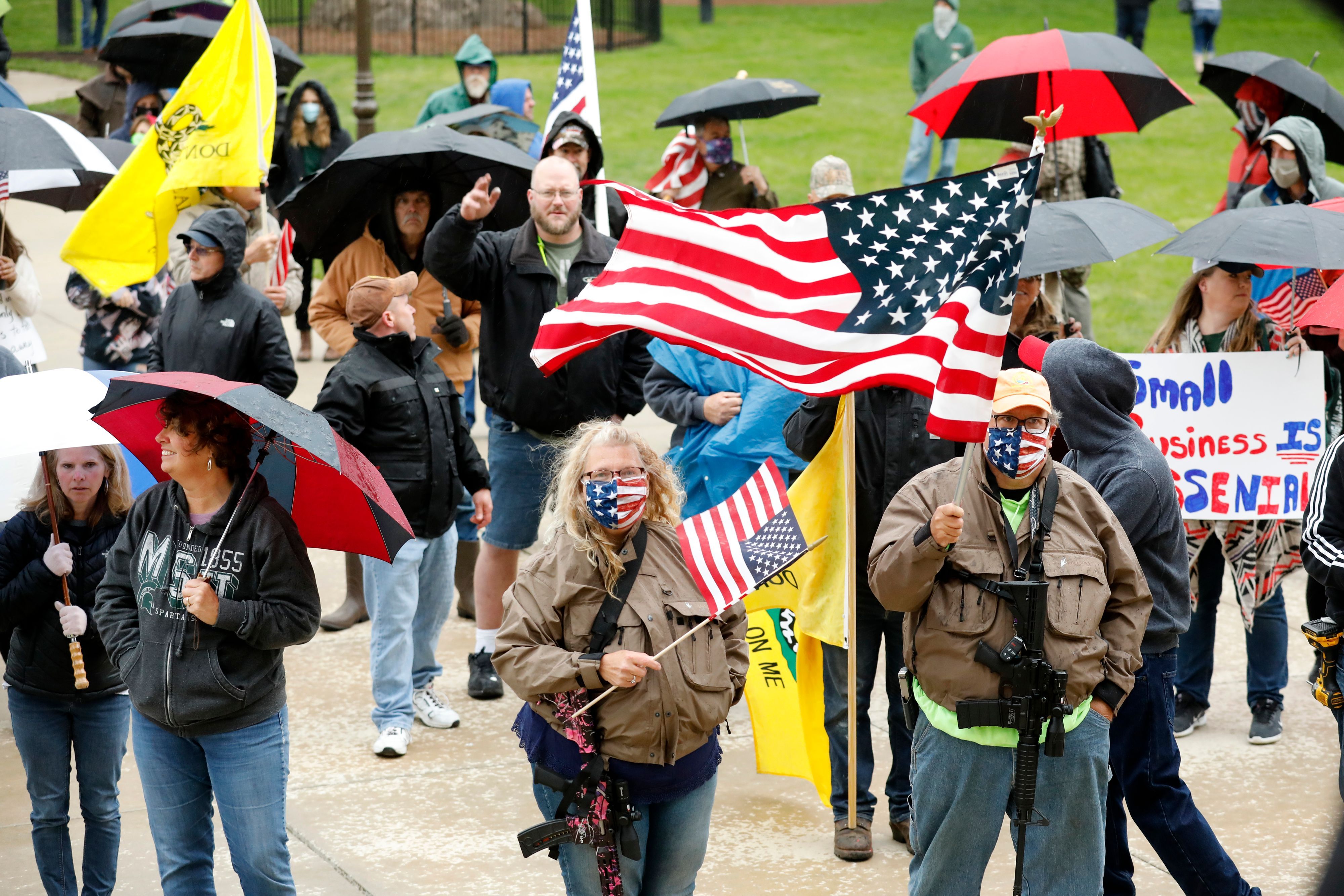 In this image, two people carry assault rifles and american flags with a crowd of people holding umbrellas standing behind them