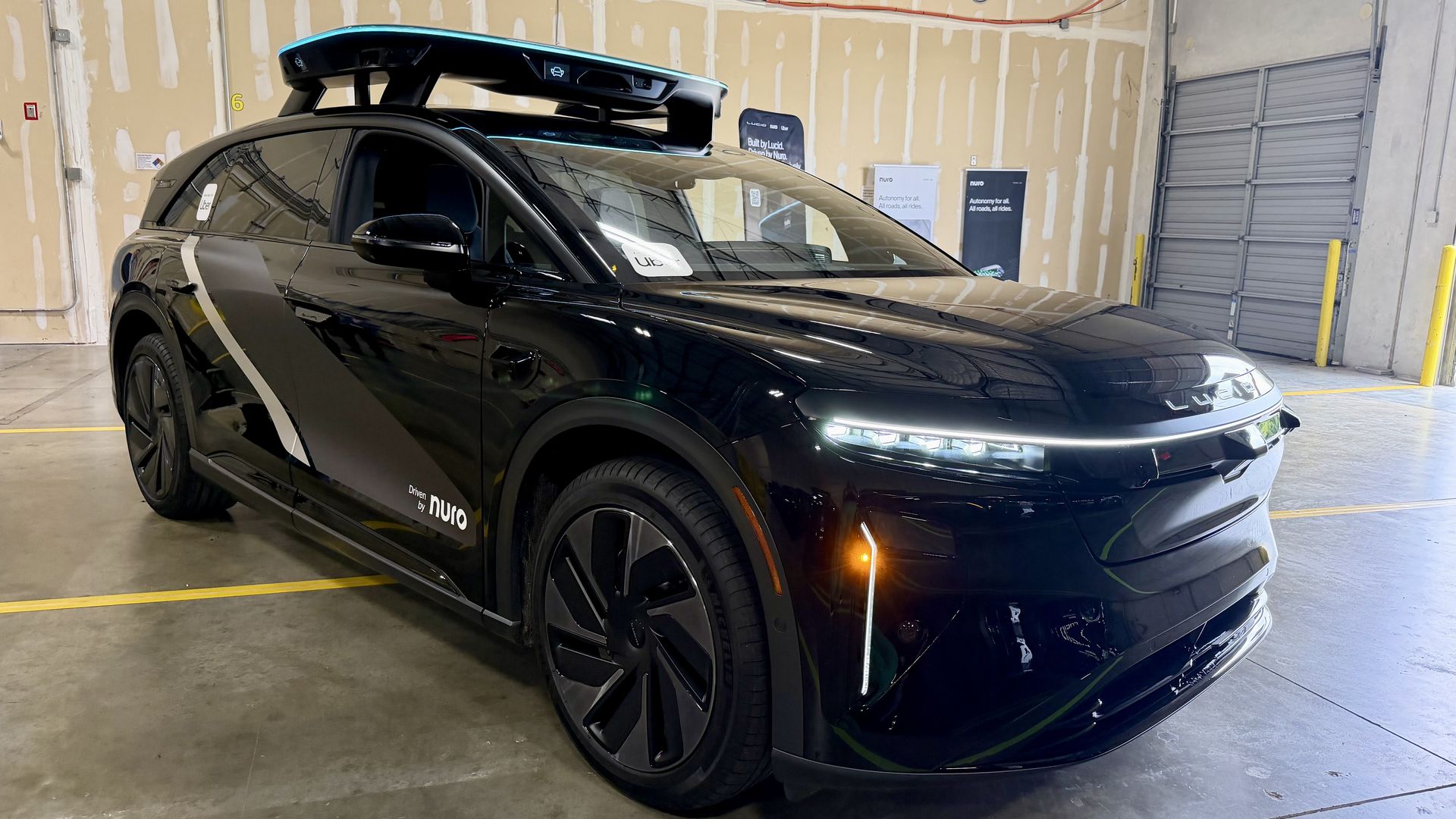 Black car with a blue-lit, rooftop sensor pod on a raised rack inside a warehouse; unfinished drywall, exposed wiring, and a "Driven by Nuro" decal on the side.