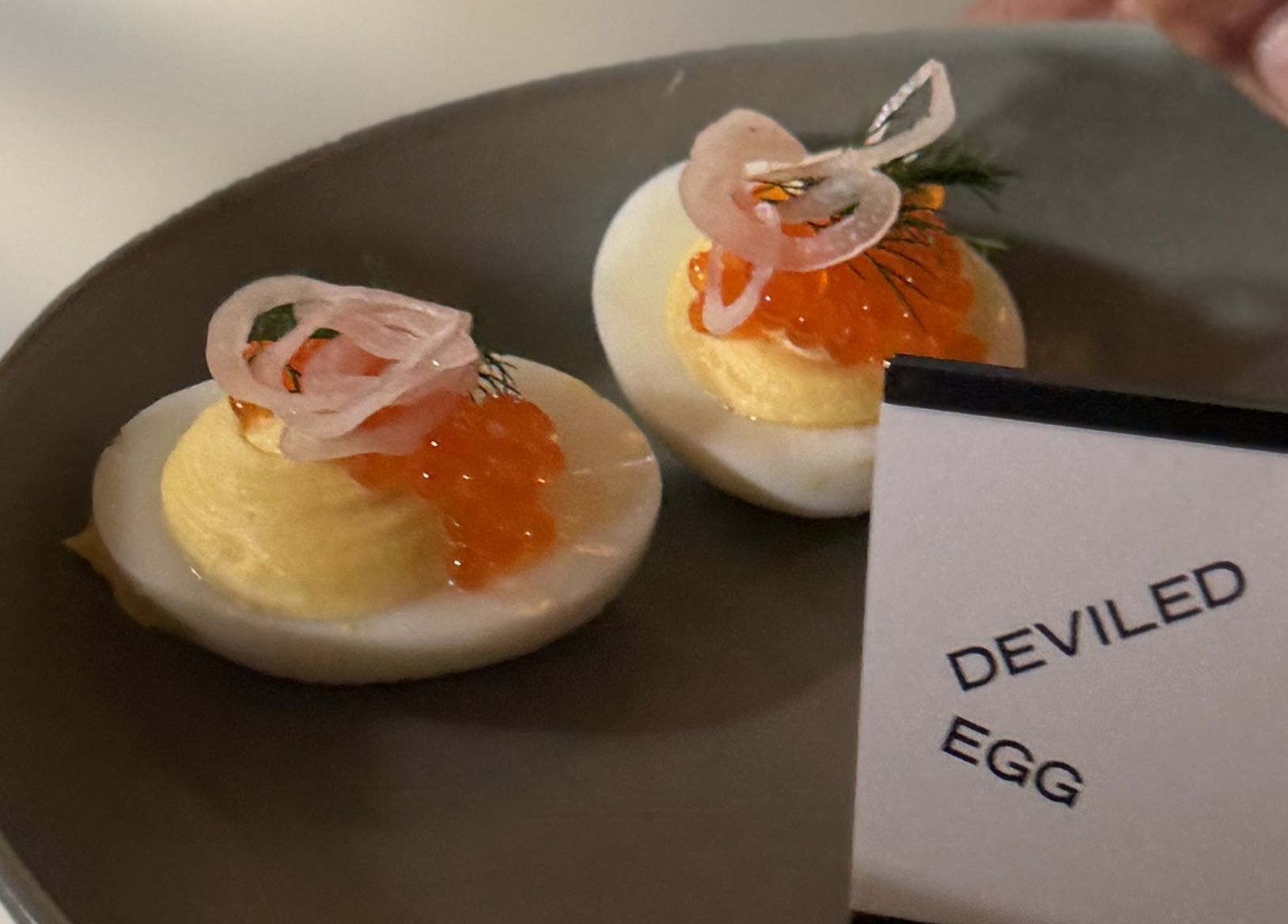 Two deviled eggs on a dark plate, pale yellow yolk filling topped with bright orange roe and a pink curled garnish; a card beside reads DEVILED EGG.