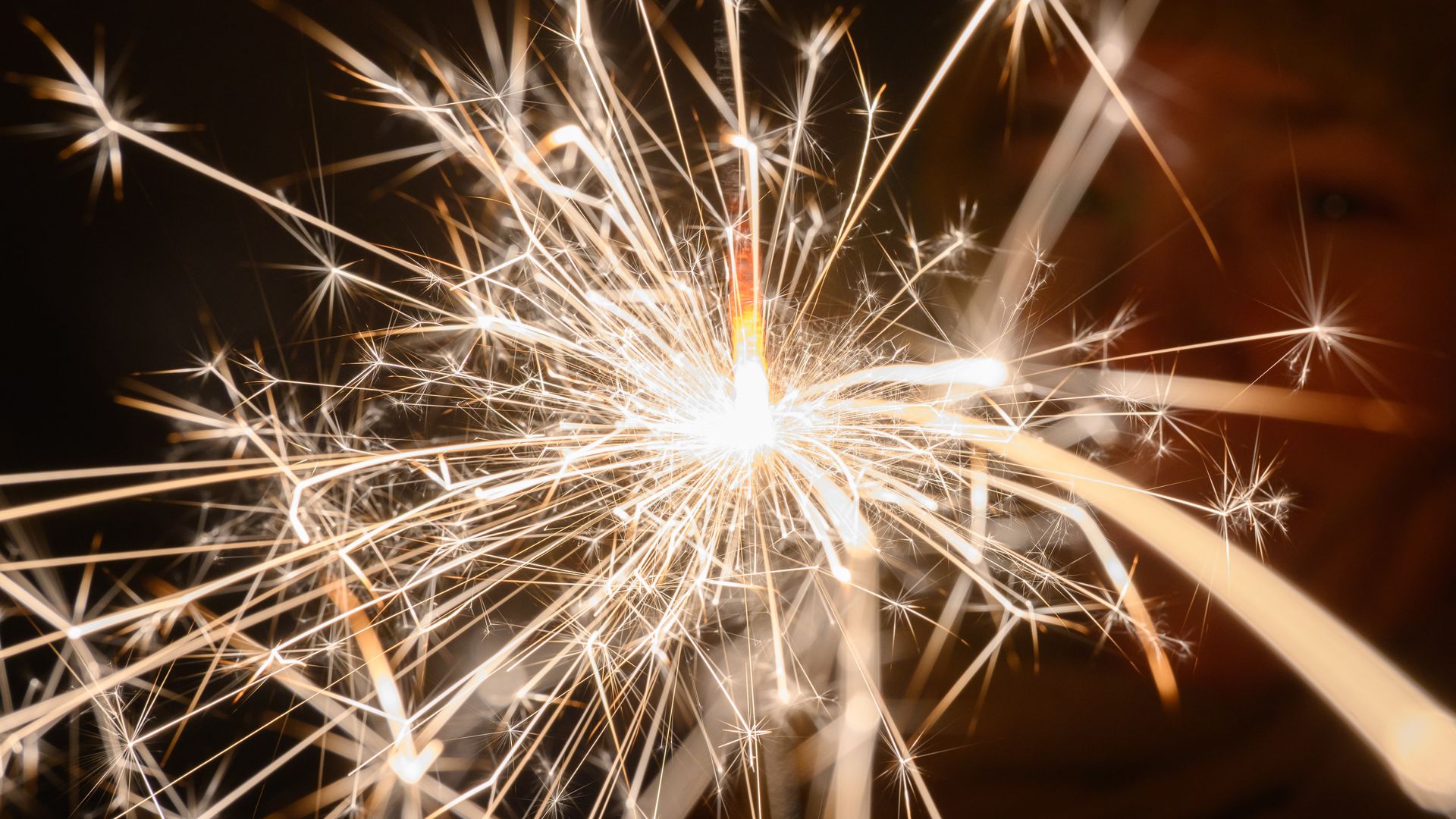 Close-up of a lit sparkler with bright, radiating sparks and a glowing orange core, set against a dark background.