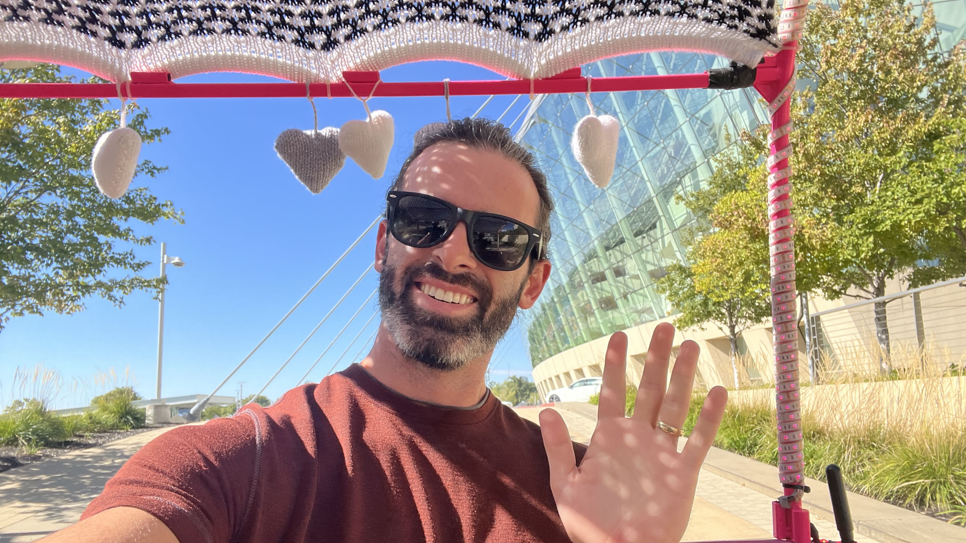 Reporter Travis smiling with sunglasses waving, wearing a maroon shirt, sitting in a pedicab under a decorated canopy with hanging hearts near the Kauffman Center on a sunny day.