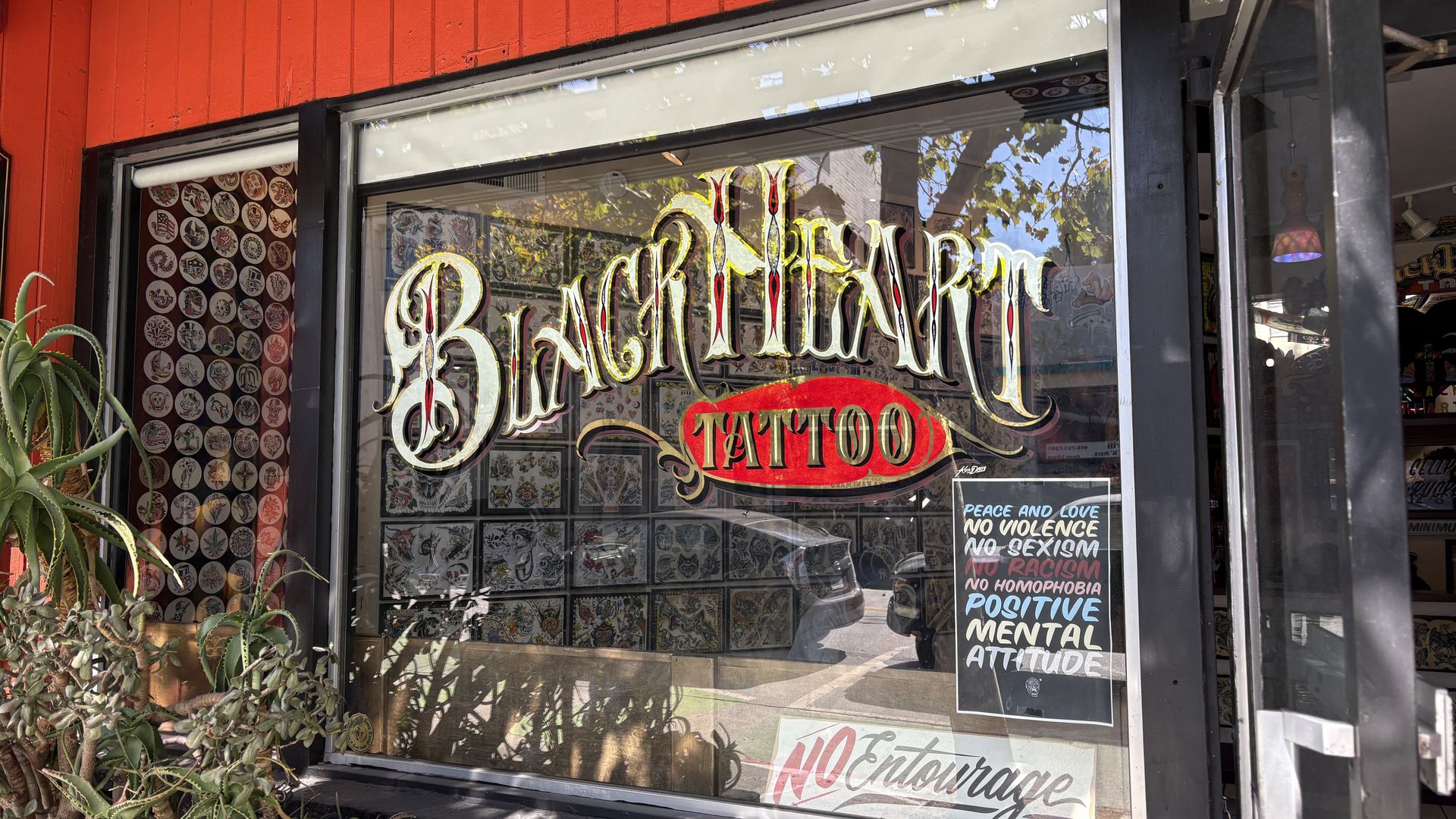 Tattoo shop window with ornate gold lettering reading "Black Market" and a red "TATTOO" banner; tattoo flash art fills the panes, red storefront, and a potted aloe plant on the left.