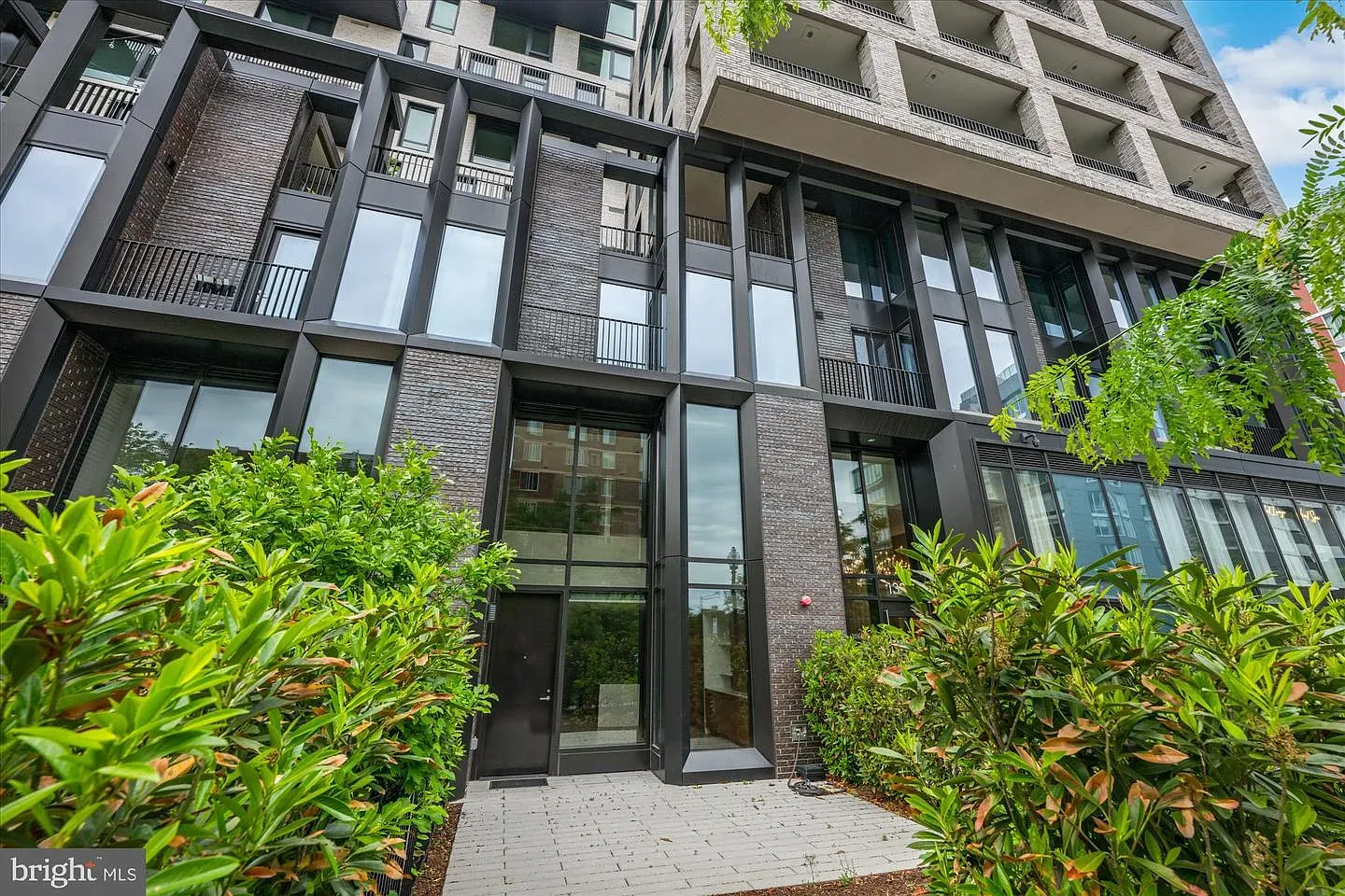 Modern brick and glass apartment building with tall black-framed windows, balconies, and lush green shrubs along a paved entrance courtyard.