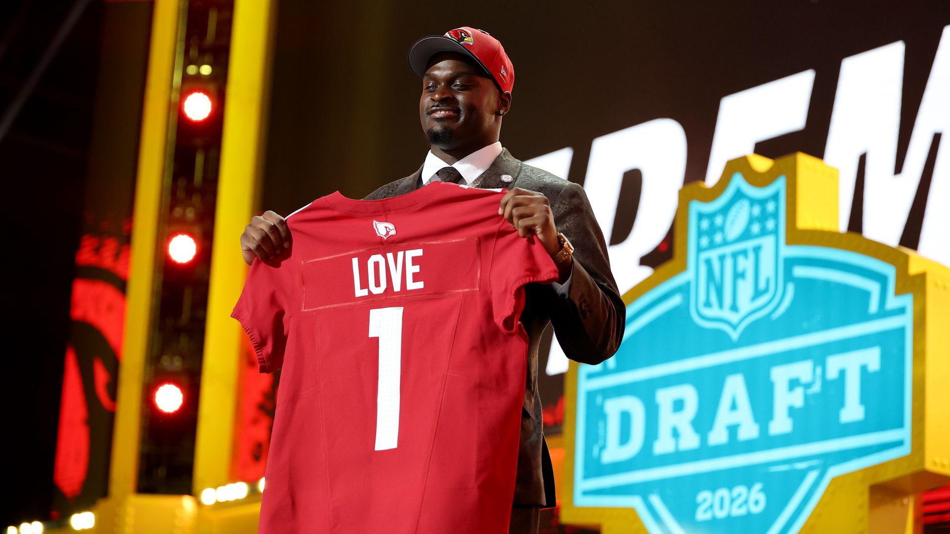 A man in a dark suit holds up a red football jersey with an Arizona Cardinals logo, the word "Love" and the number 1 on it, while wearing a red Arizona Cardinals hat. A light blue NFL logo with the words "Draft 2026" under it are behind him.