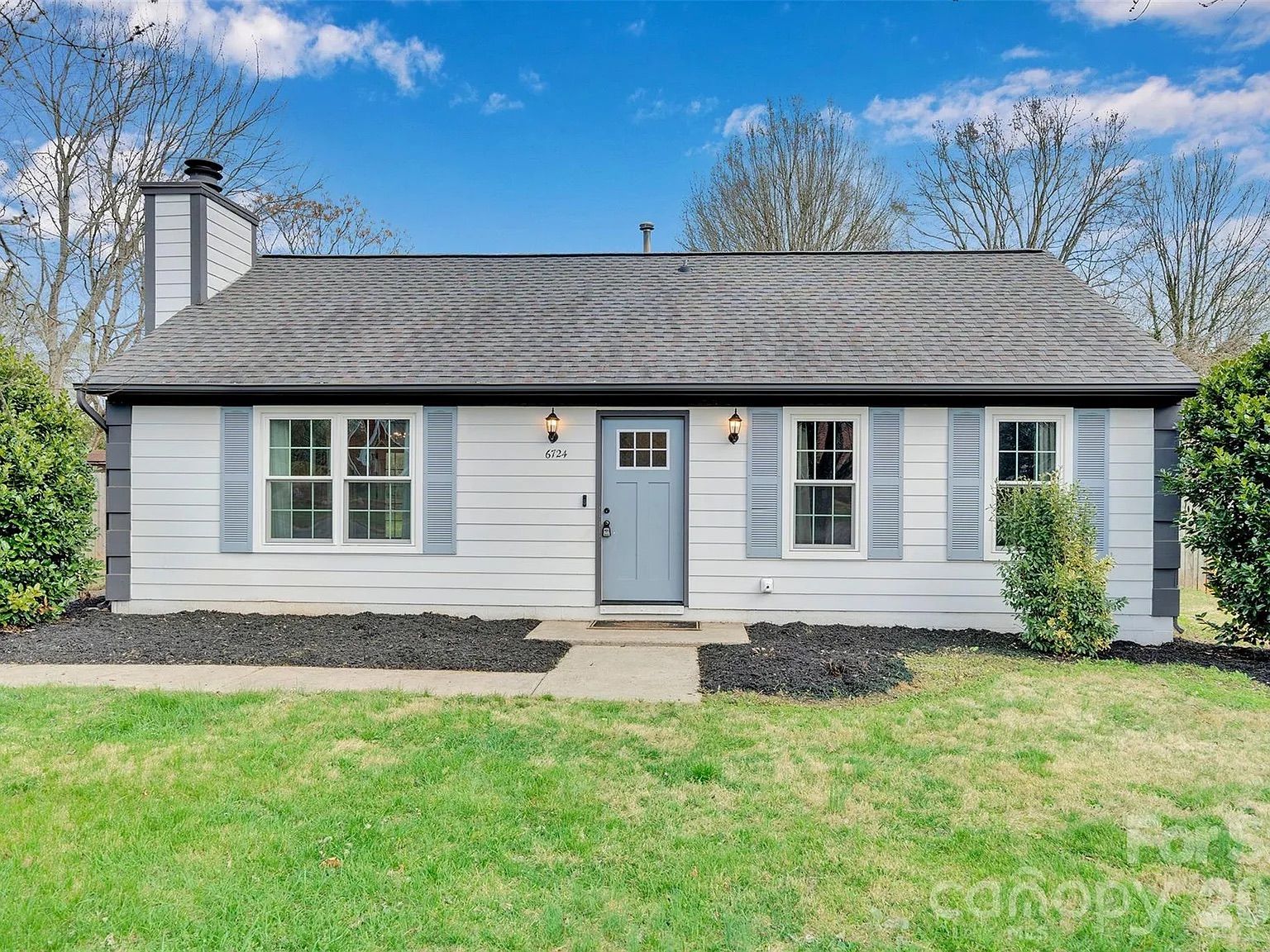 Single-story ranch home with light gray siding, blue shutters and front door, black roof, two lantern lights by the entry, mulched beds, green lawn, leafless trees, and a bright blue sky.