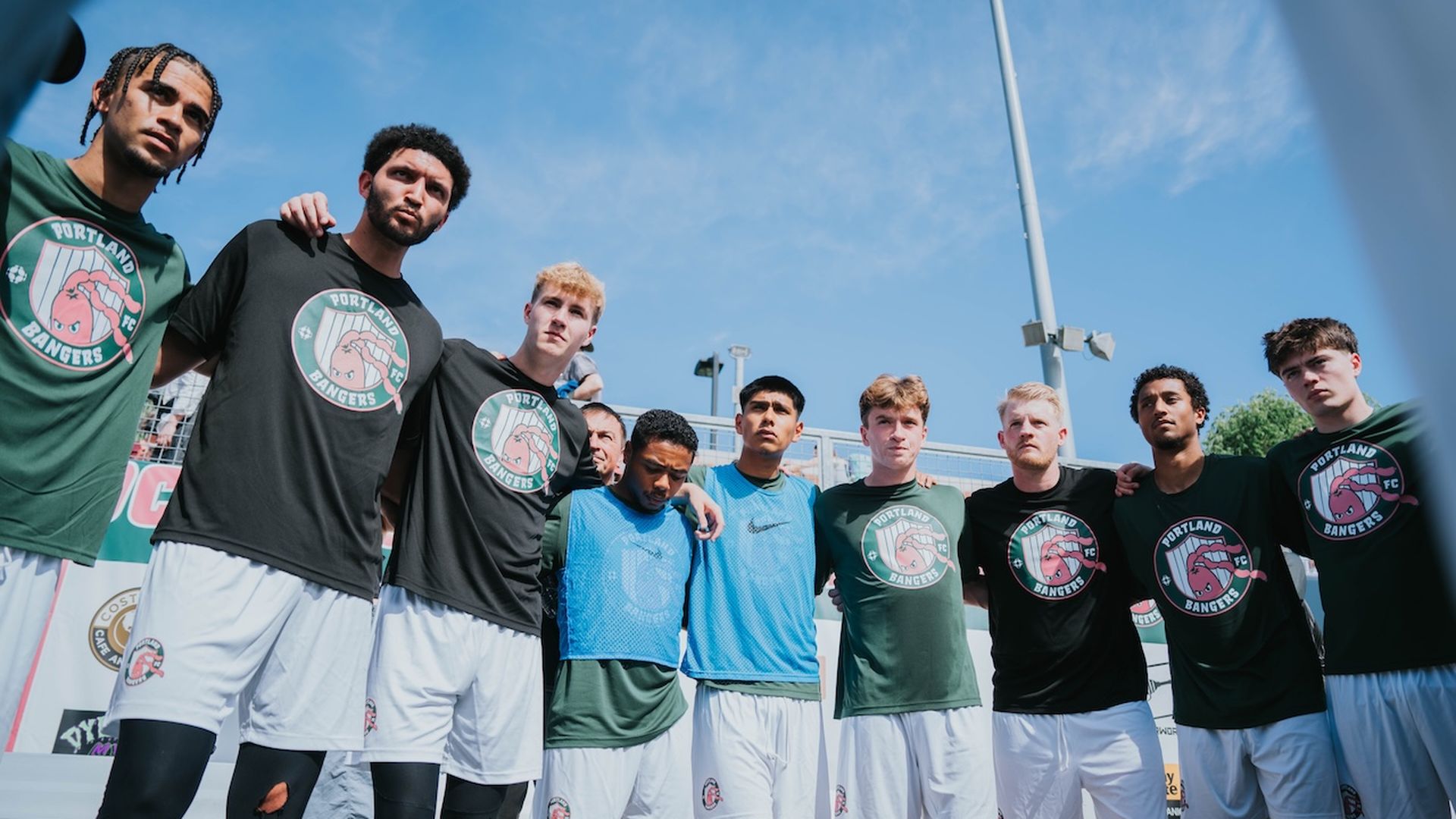 The Portland Bangers soccer team stands arm-in-arm in a pregame huddle, wearing branded shirts and shorts, showing unity and focus before their debut match under a clear blue sky.