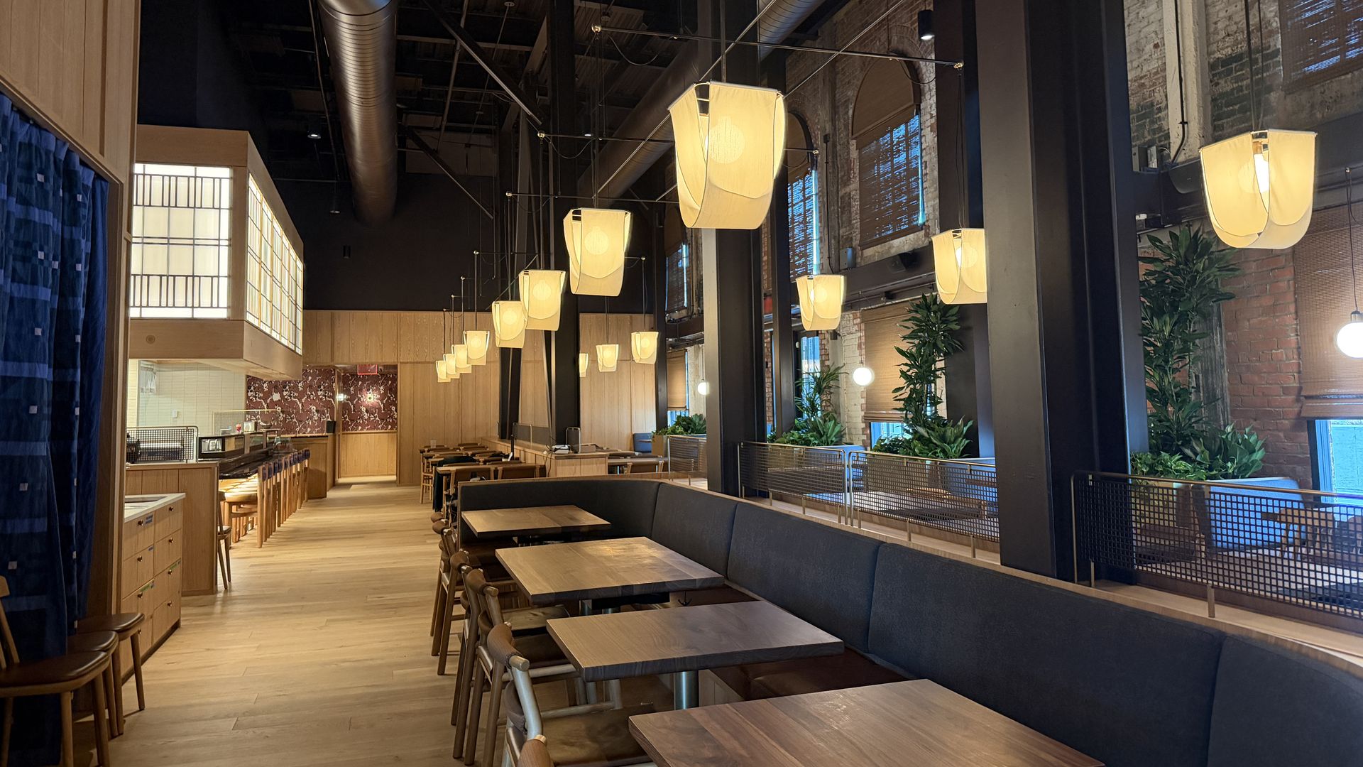 Interior of a modern restaurant with wooden tables and chairs, hanging yellow lantern-style lights, black ceiling with exposed ducts, brick walls, and large windows with plants on the ledge.