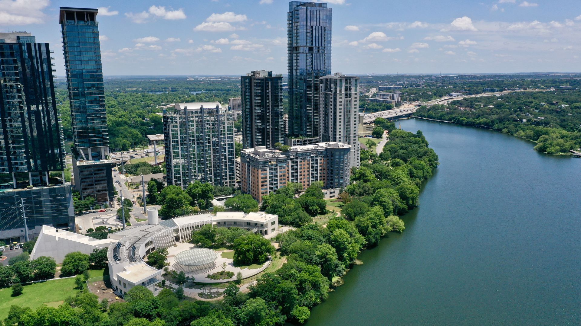 Aerial view of a modern city with tall blue glass skyscrapers along a winding river, green parks, a bridge, and highways stretching into the distance under a bright blue sky with scattered clouds.