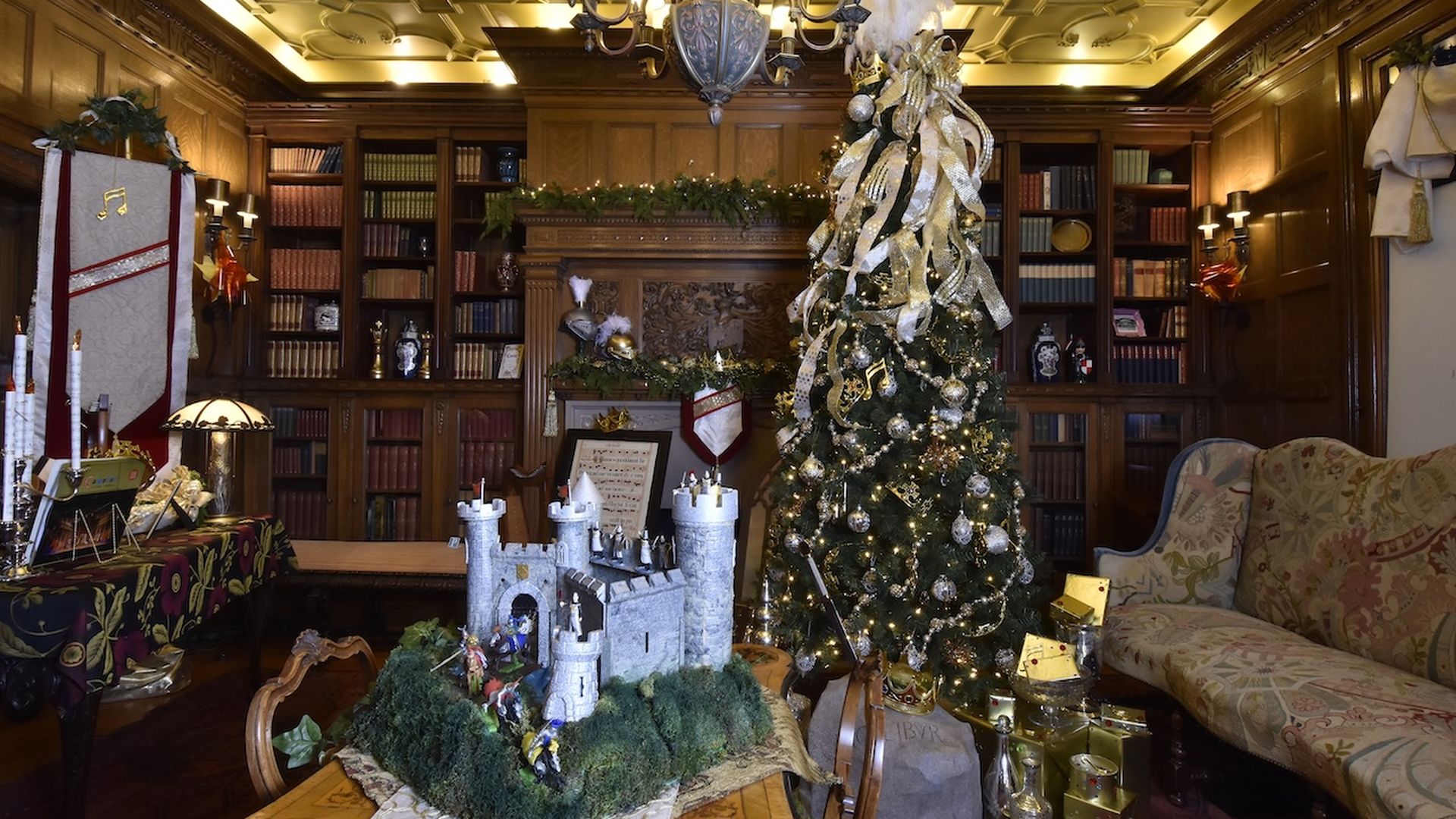 Cozy library room with wooden shelves and a decorated Christmas tree with gold ribbons and ornaments, a medieval castle model on a table, and festive wrapped gifts nearby.