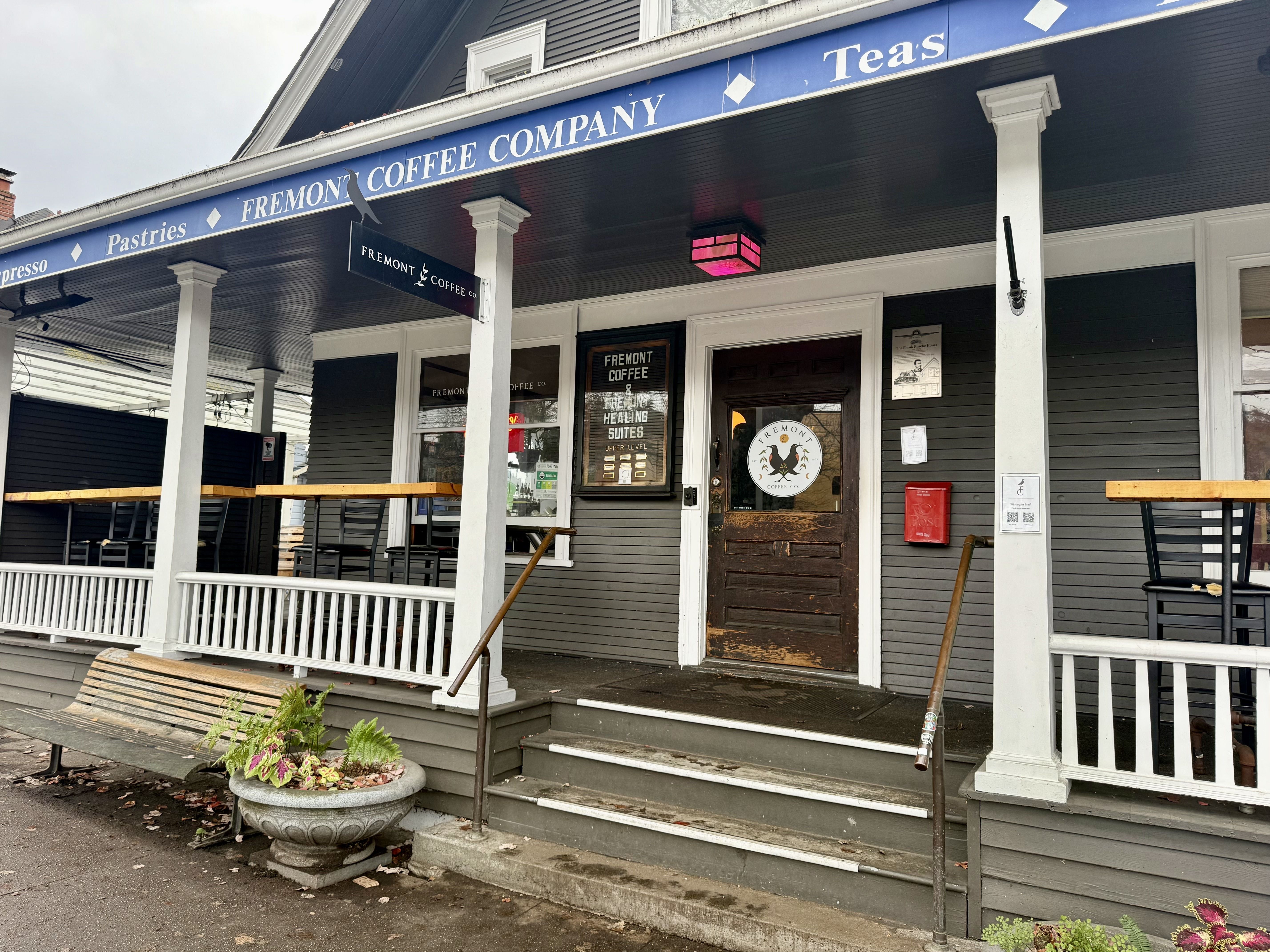 Front porch of Fremont Coffee Company with gray siding, white columns, wooden bench, plants in a stone planter, and a dark wooden door featuring the coffee company logo.