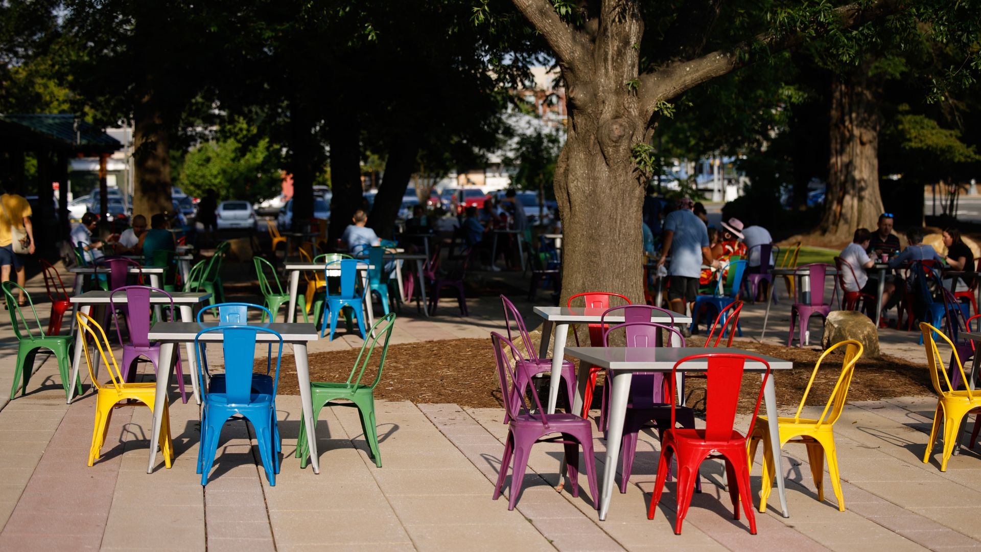 Outdoor seating area with colorful metal chairs in blue, yellow, red, purple, and green around gray tables under large trees with people sitting and socializing in the shaded background.