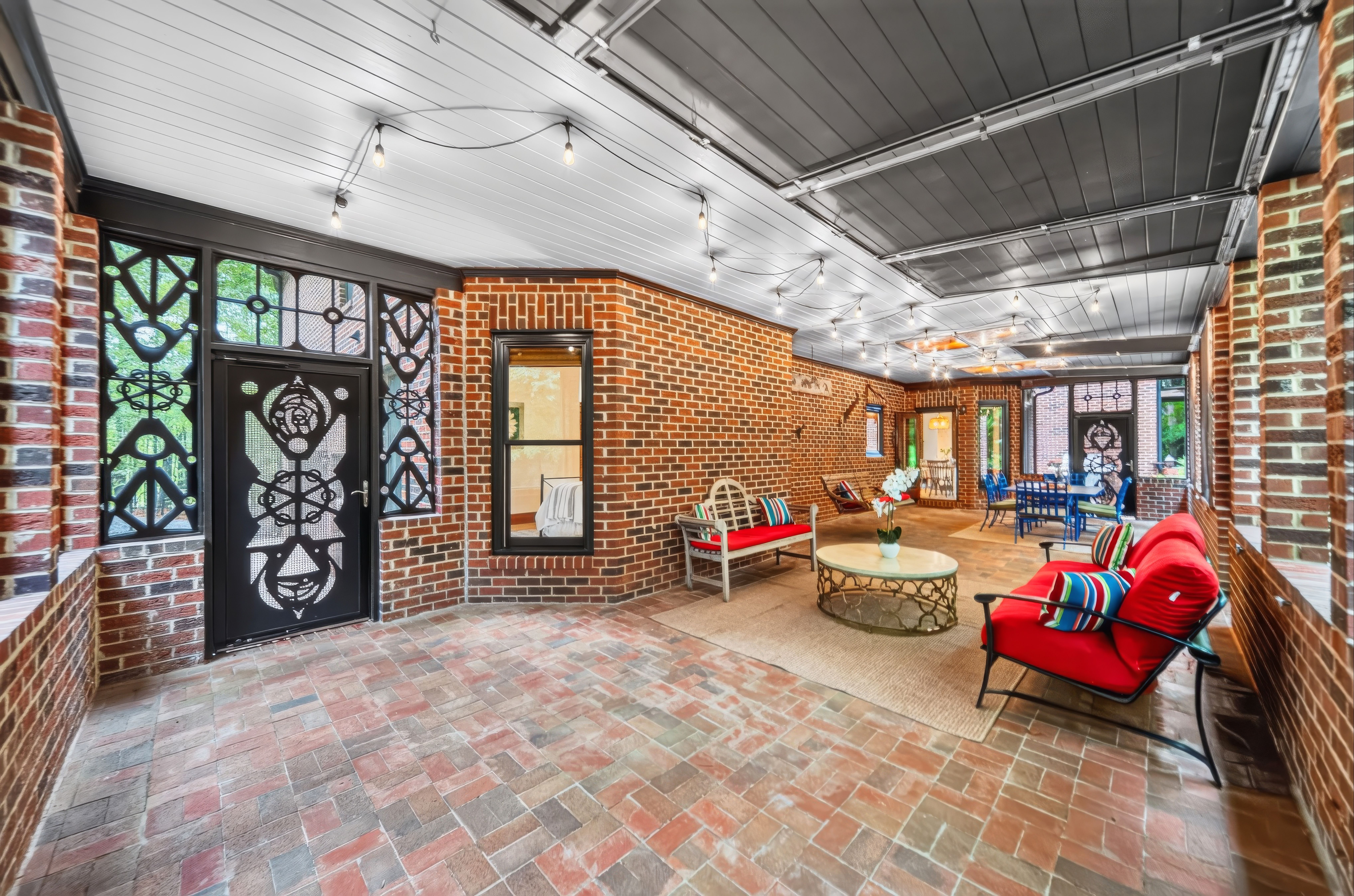 Covered brick patio with string lights, a gray ceiling, and brick walls. Black ornate metal doors, red cushioned seating, blue chairs, and round coffee tables create a cozy outdoor living area.