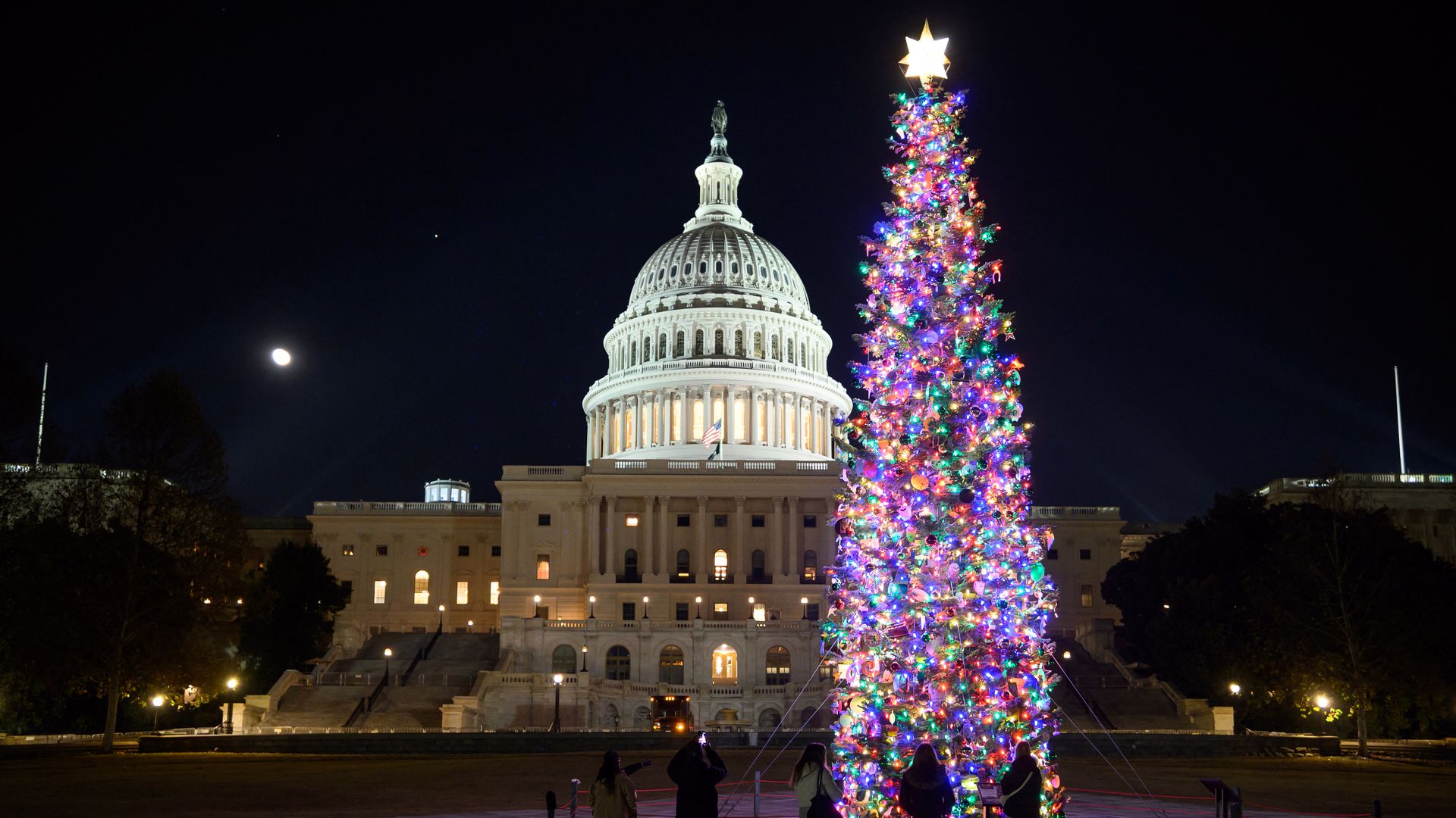 Nighttime view of the U.S. Capitol building with a large, brightly lit Christmas tree decorated with colorful lights and a glowing star on top; four people stand nearby admiring it.
