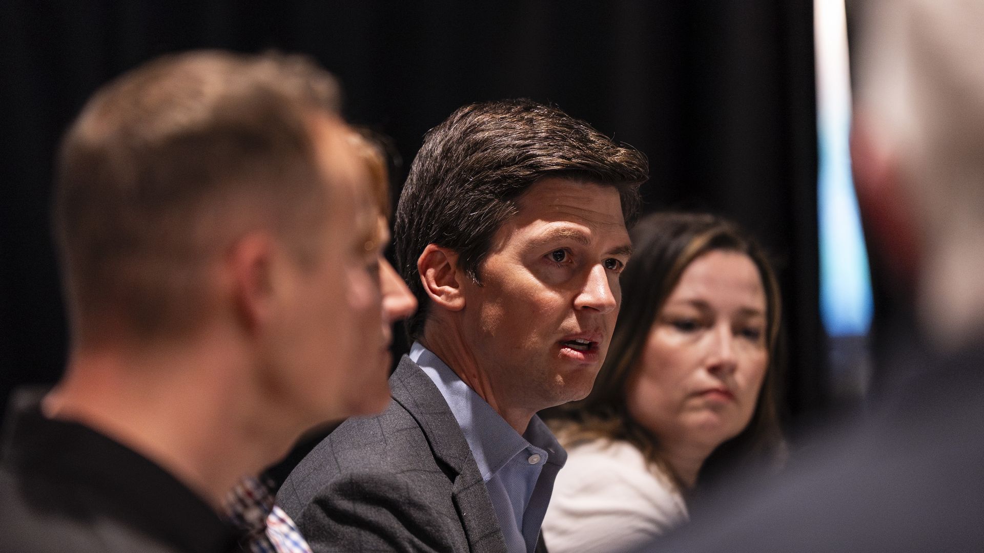 A man with dark hair wearing a gray suit and light blue shirt sits at a panel, looking to the right. Blurred faces of other attendees flank him, against a dark backdrop.