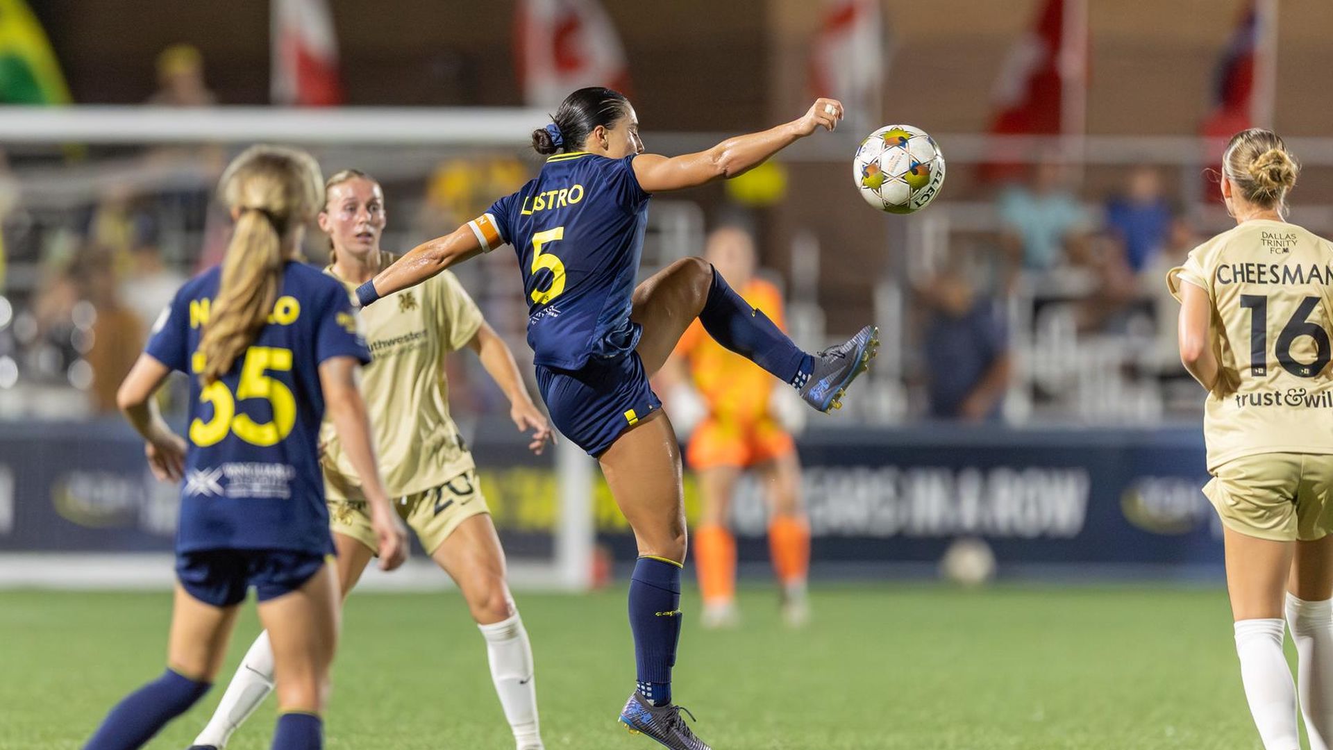 Women’s soccer game: blue jersey player #5 leaps to kick the ball midair while beige-clad opponents watch, on green turf with flags in the background.
