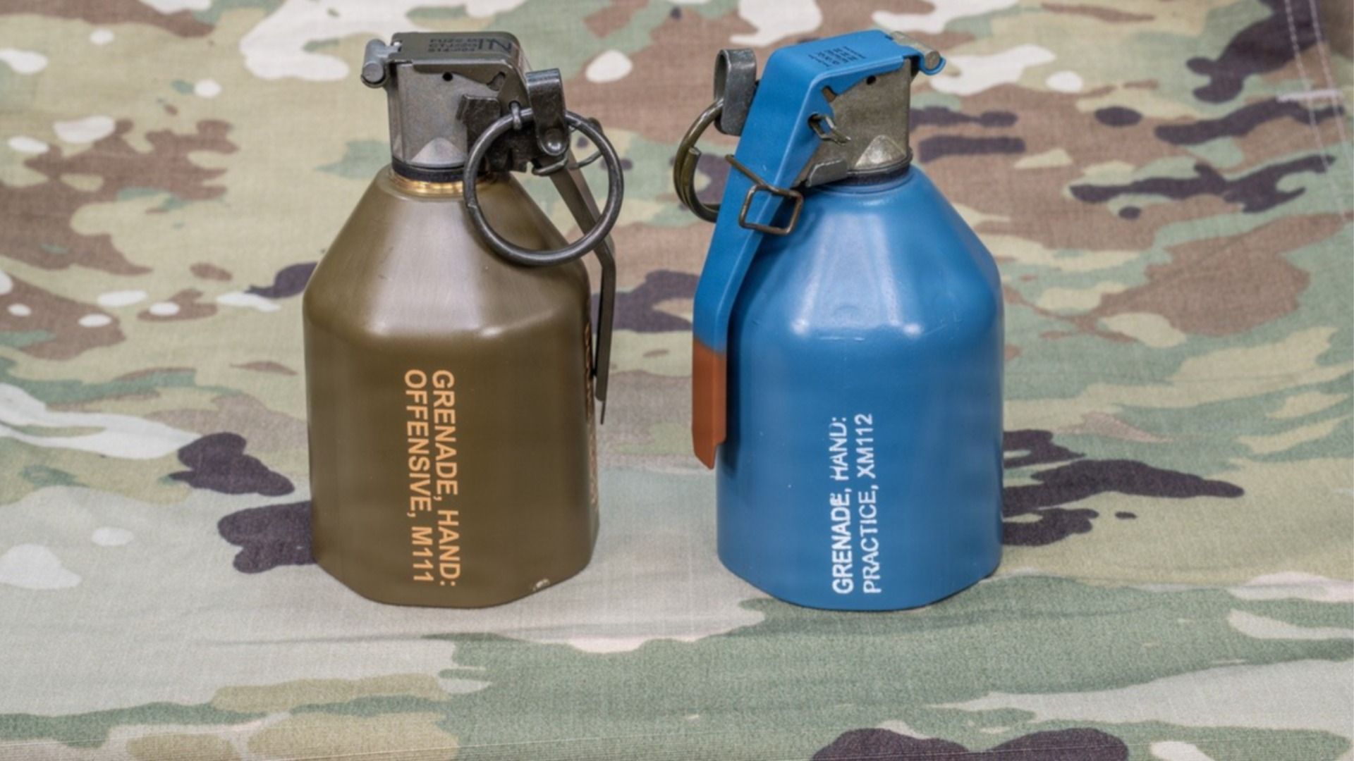 Two hand grenades, olive green and blue, with safety pins sit on camouflage fabric.