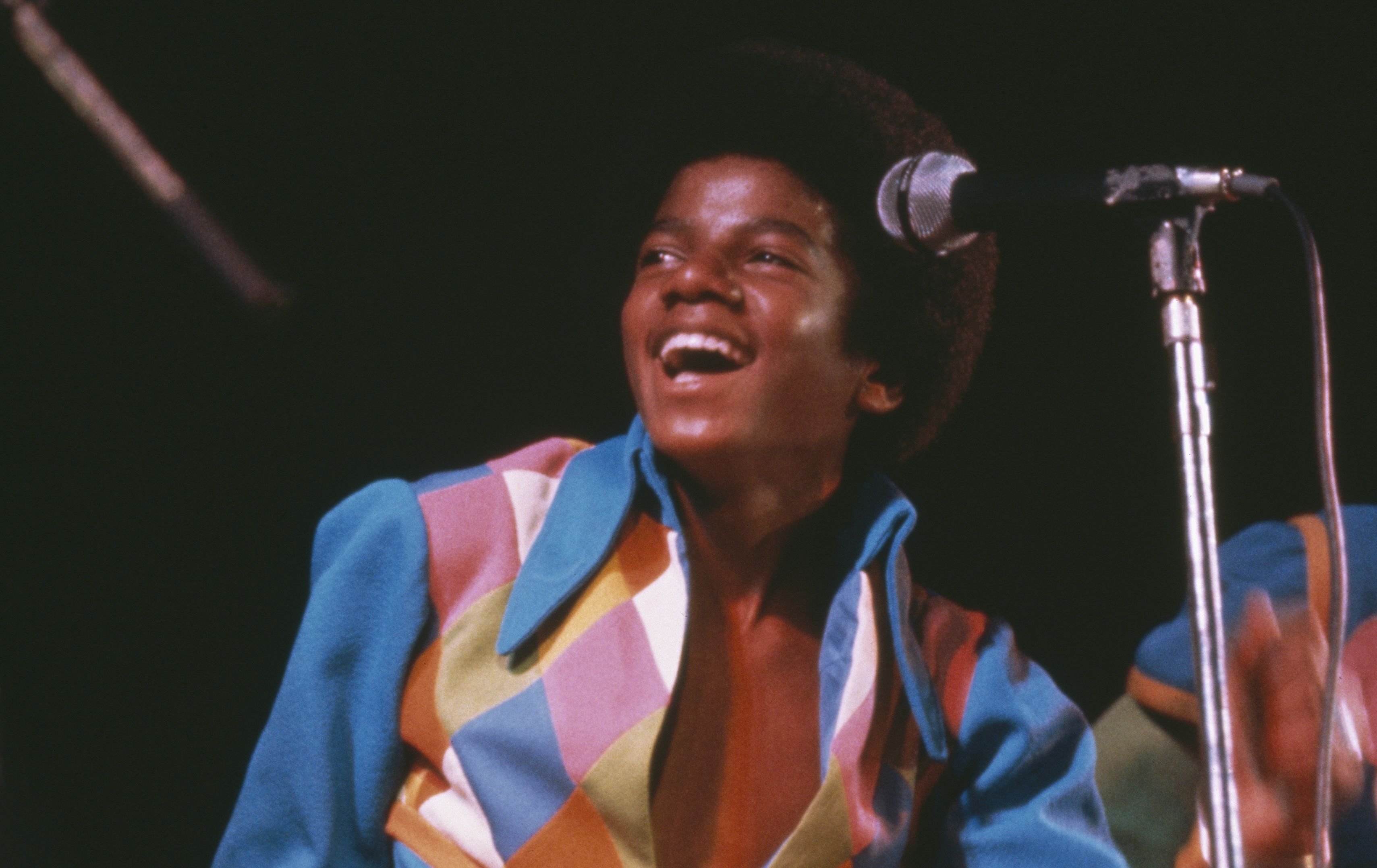 A joyful Black male singer with an Afro on stage, wearing a bright, multicolored striped shirt, singing into a microphone on a stand against a dark background.