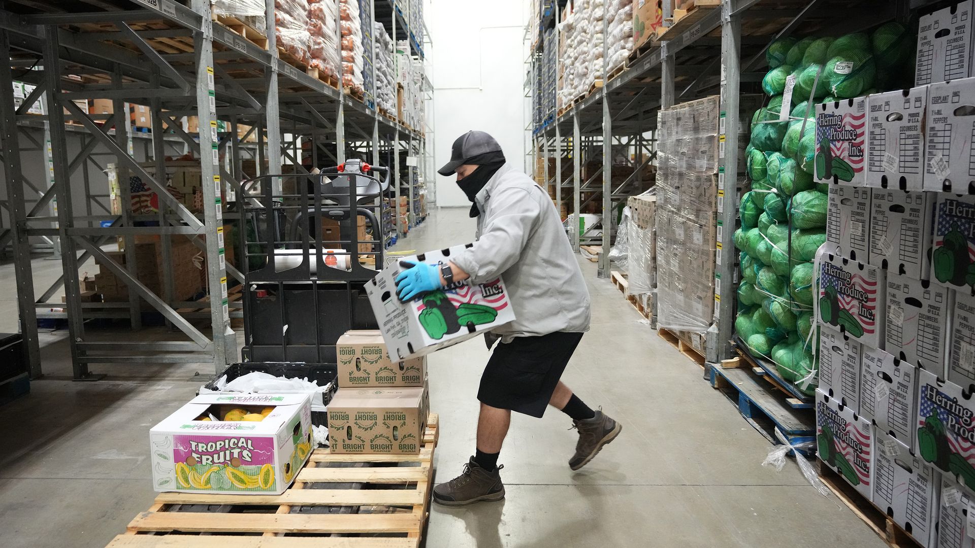 A worker in a gray jacket, cap and face mask moves through a tall warehouse aisle, lifting a Tropical Fruits box from a pallet while shelves are stacked with cardboard boxes and green sacks.
