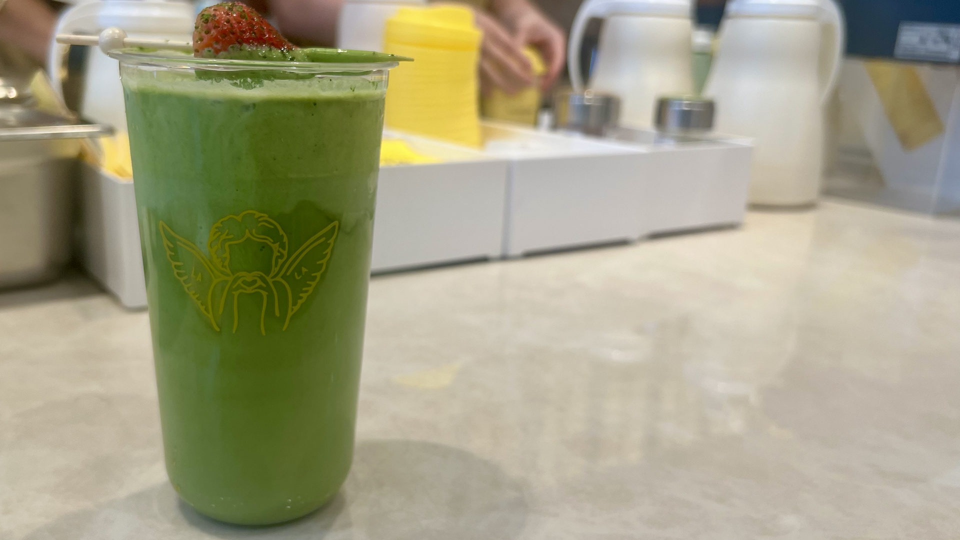 Green drink in a transparent cup with a yellow angel logo, topped with a strawberry slice, placed on a light-colored counter with blurred background of condiment containers and hands.