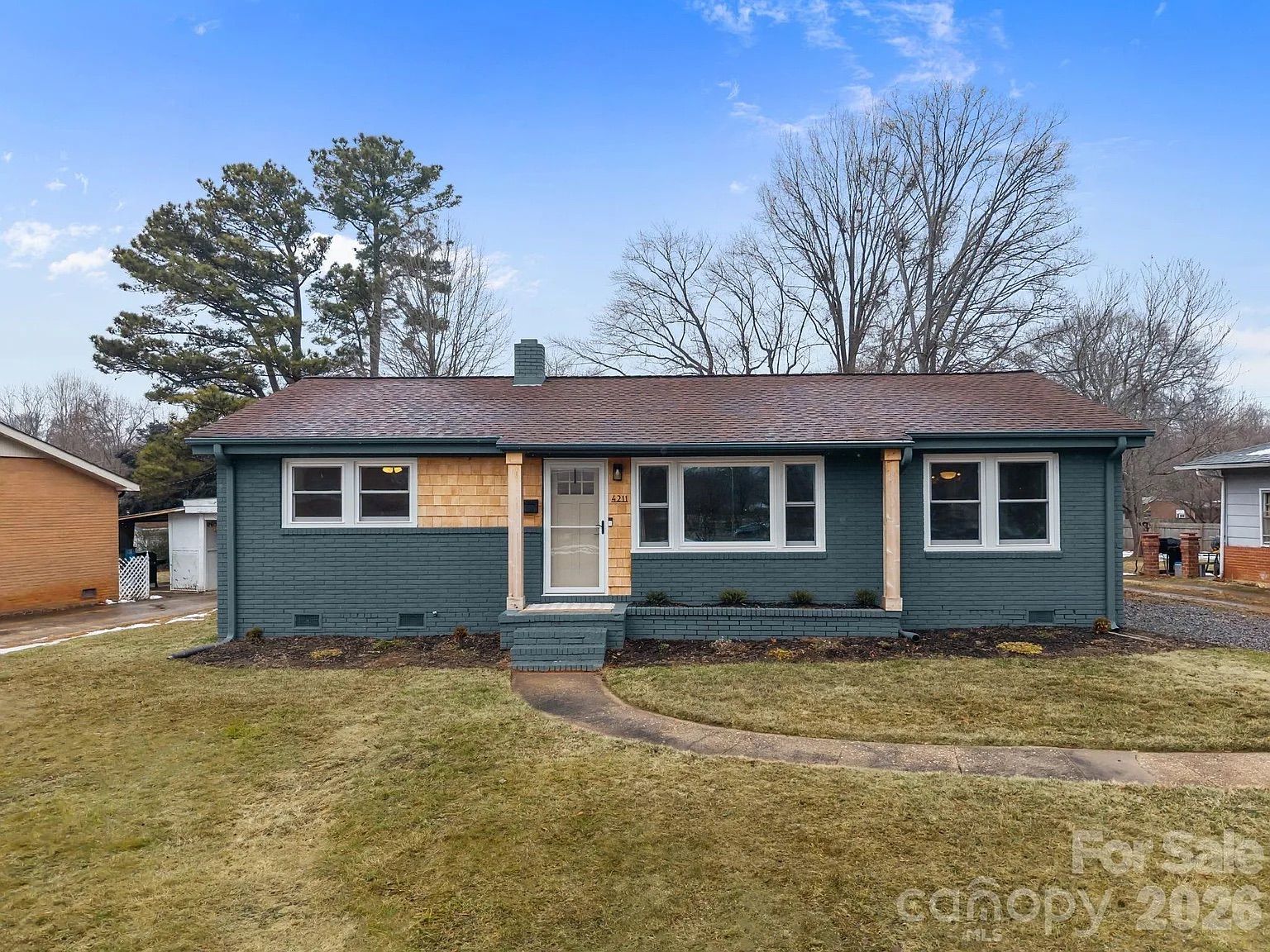 Single-story house with dark green brick, tan upper half, brown roof, white door, and windows, set against a partly cloudy sky and leafless trees, with a front yard and curved path.