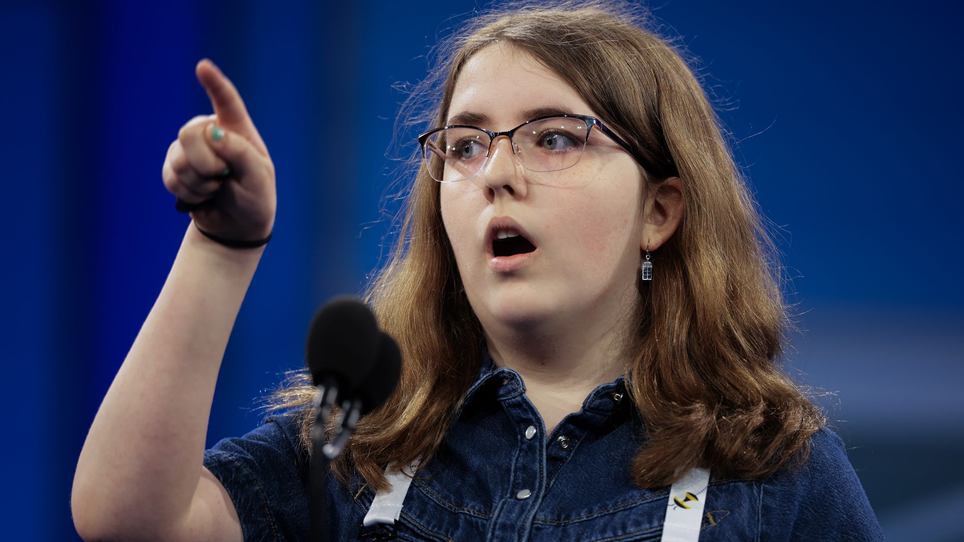 Young girl in blue polo shirt and glasses behind a microphone, pointing her finger.