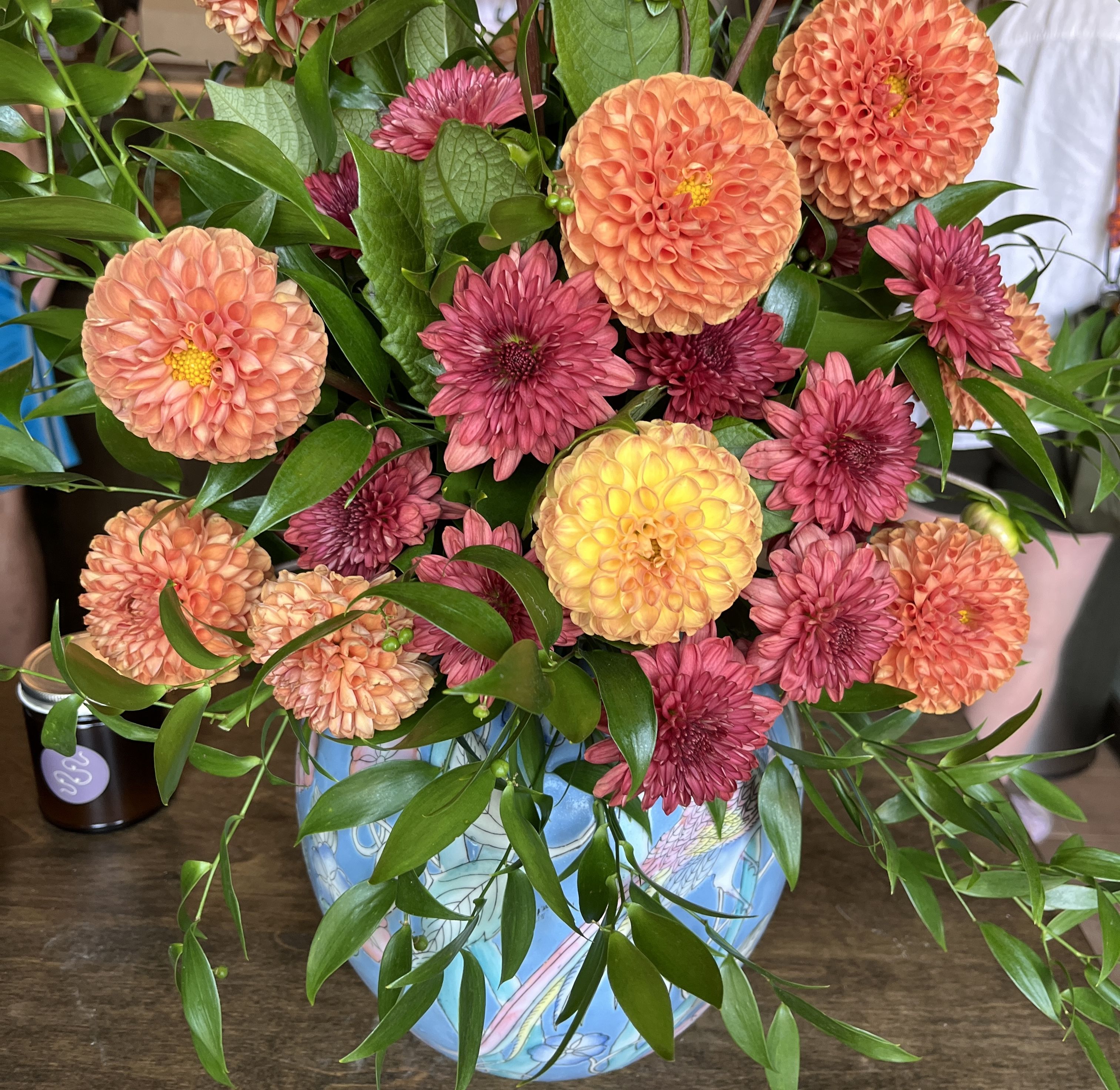 Bouquet of orange, yellow, and deep pink flowers with green leaves in a blue vase decorated with a pink bird, set on a wooden surface indoors.