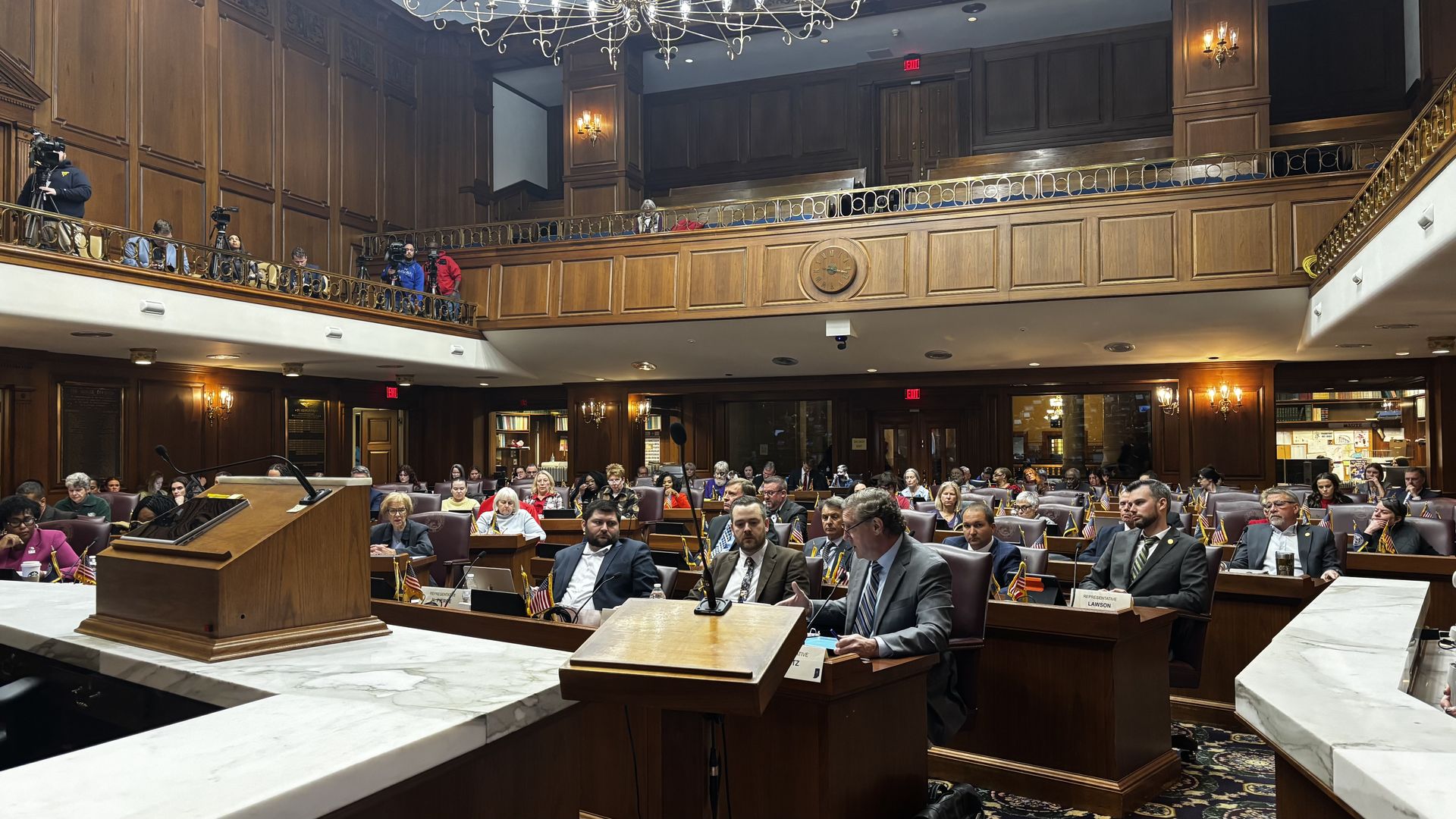 Wood-paneled legislative chamber filled with seated officials and audience, small American flags on desks, cameras on upper balcony, warm lighting, and a clock on back wall.