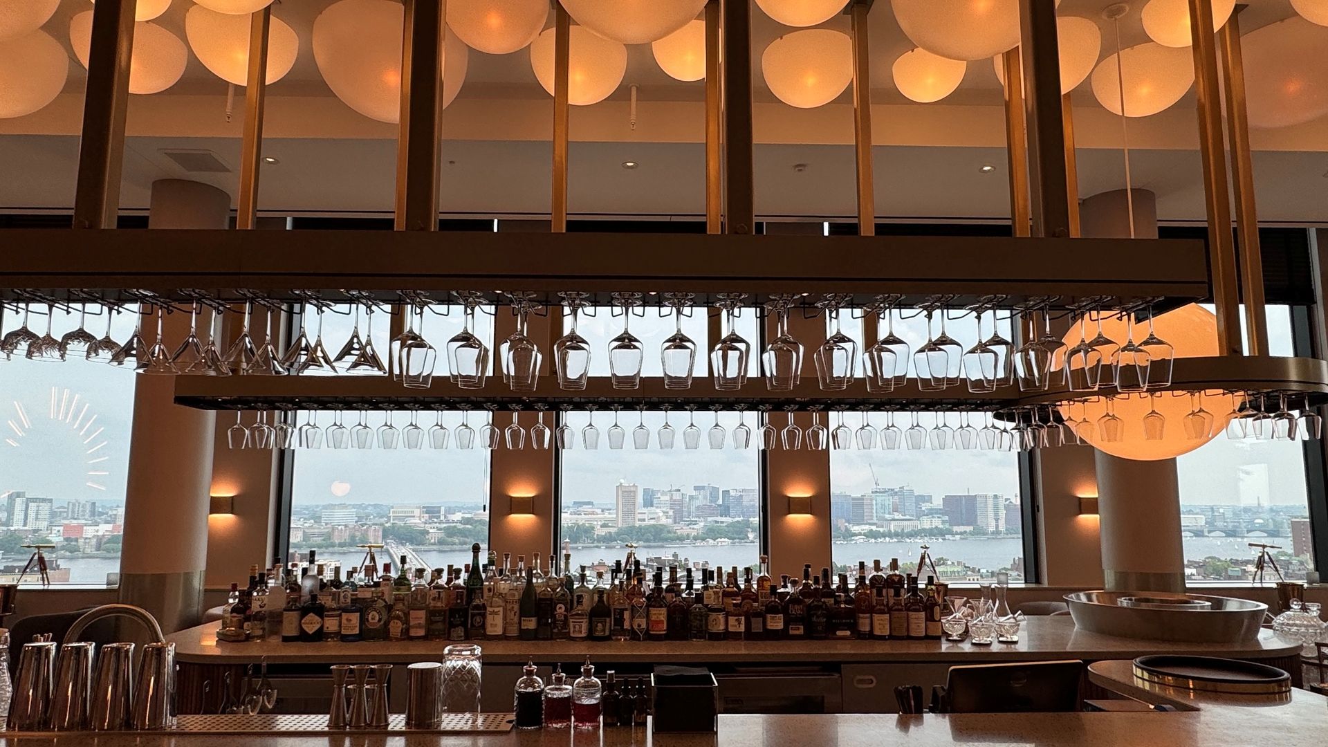 Interior view of a modern bar with hanging wine glasses, assorted bottles on the counter, orange globe lights, large windows showing a city skyline and river in the background.